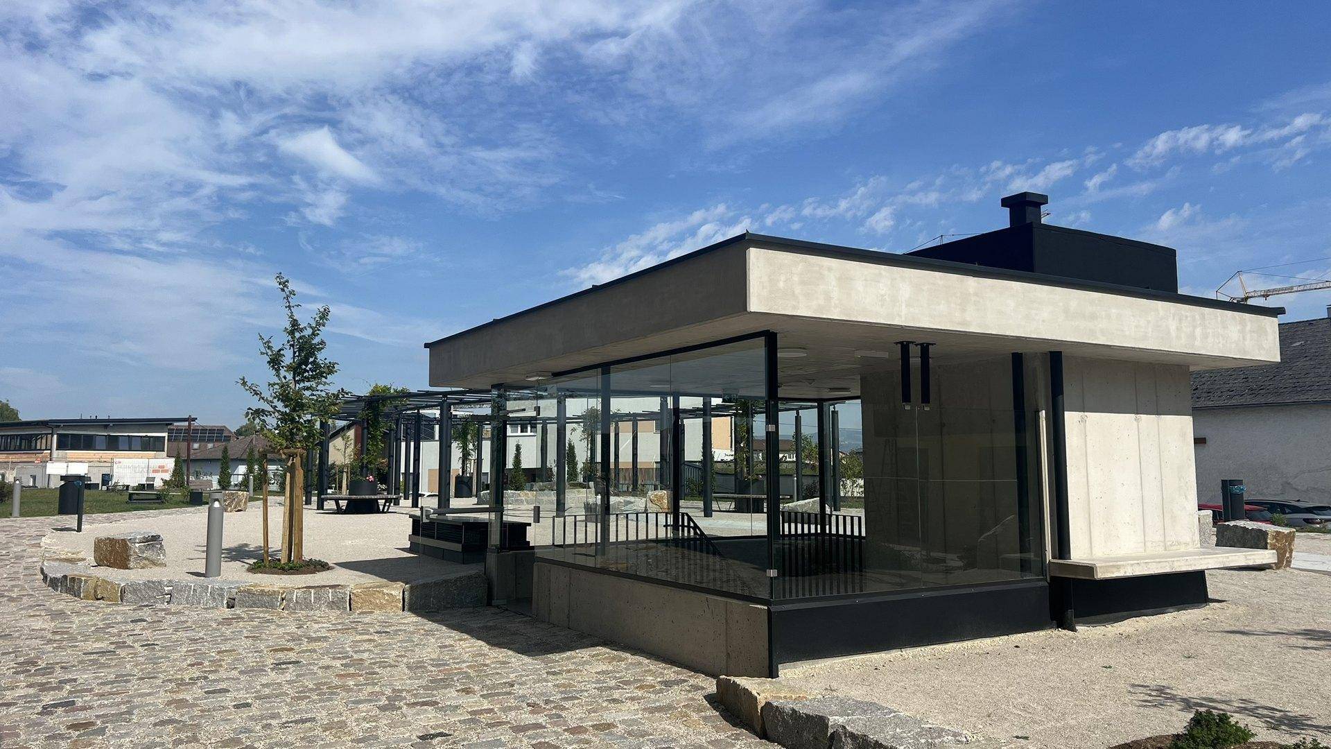 Moderner Pavillon mit Glaswänden und einem Vordach in einem weitläufigen Außenbereich.