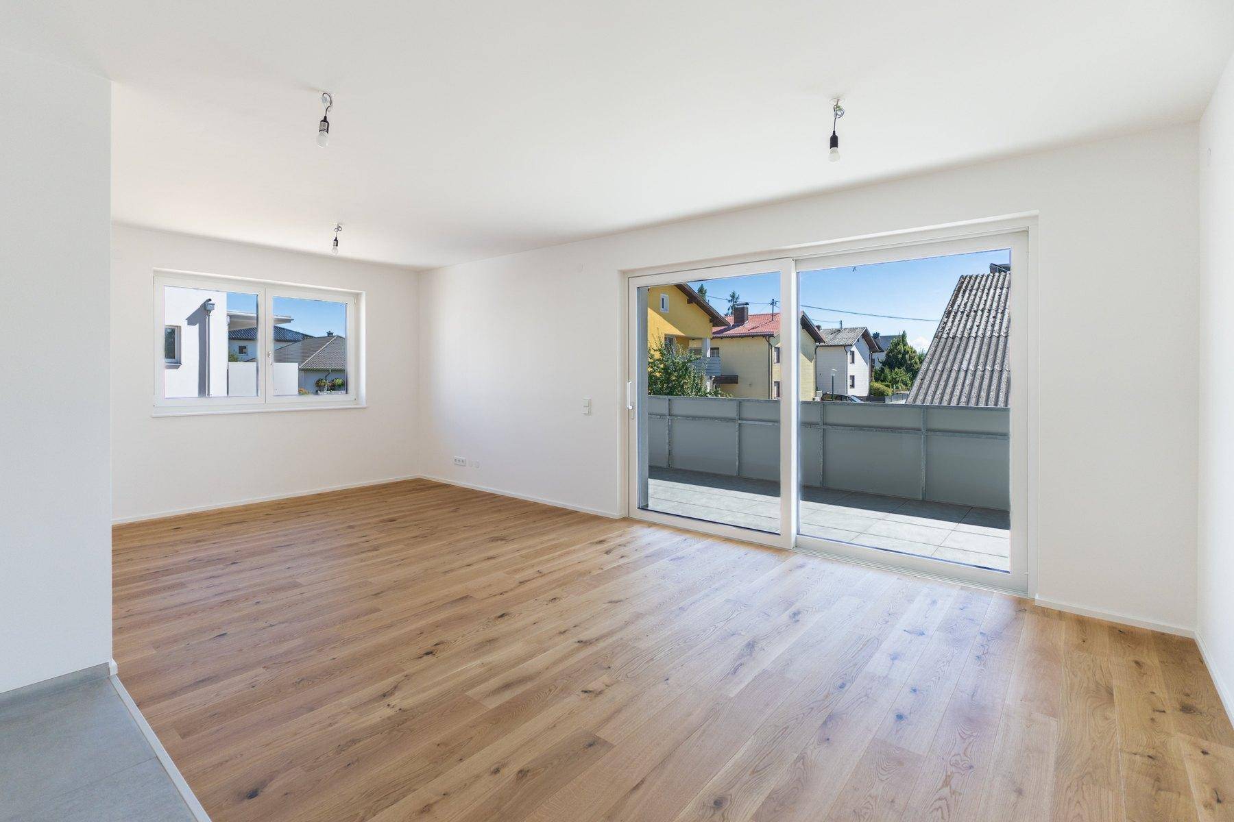 Heller, leerer Wohnraum mit hochwertigem Holzboden, großen Fenstern und Zugang zu einer Terrasse.