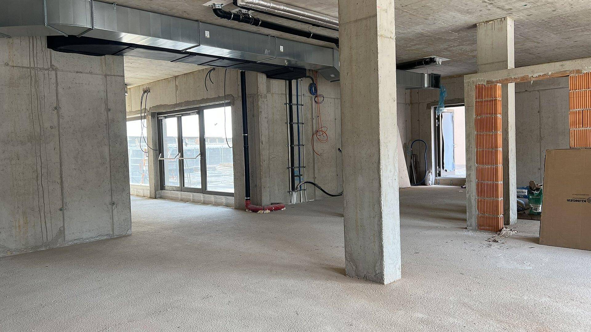 Rohbau-Innenraum mit Betonstützen, freiliegenden Installationen und großen Fenstern, die den Raum erhellen.