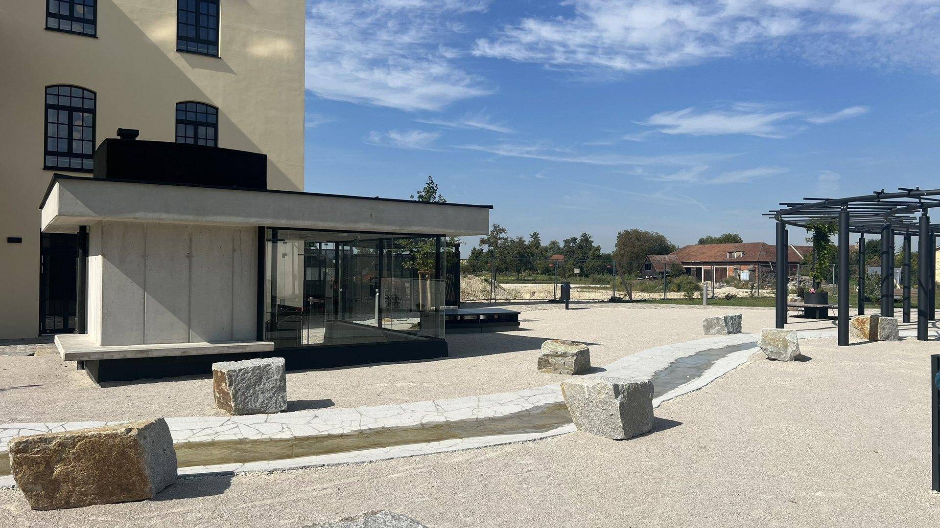 Moderner Pavillon mit Glaswänden in einem Außenbereich mit Kies und großen Steinen.