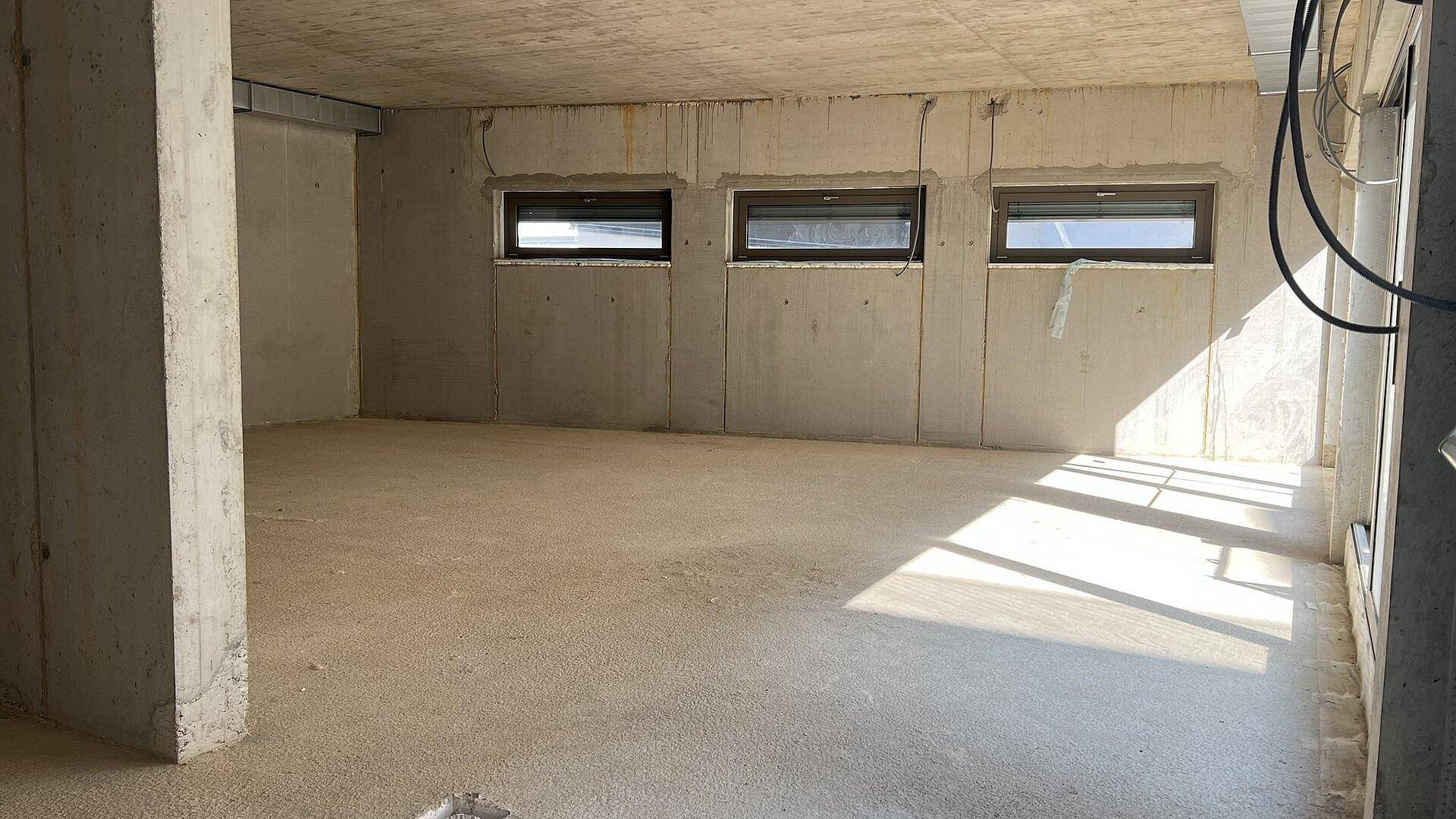 Rohbau-Raum mit Betonwänden und drei Fenstern, die Sonnenlicht auf den Betonboden werfen.