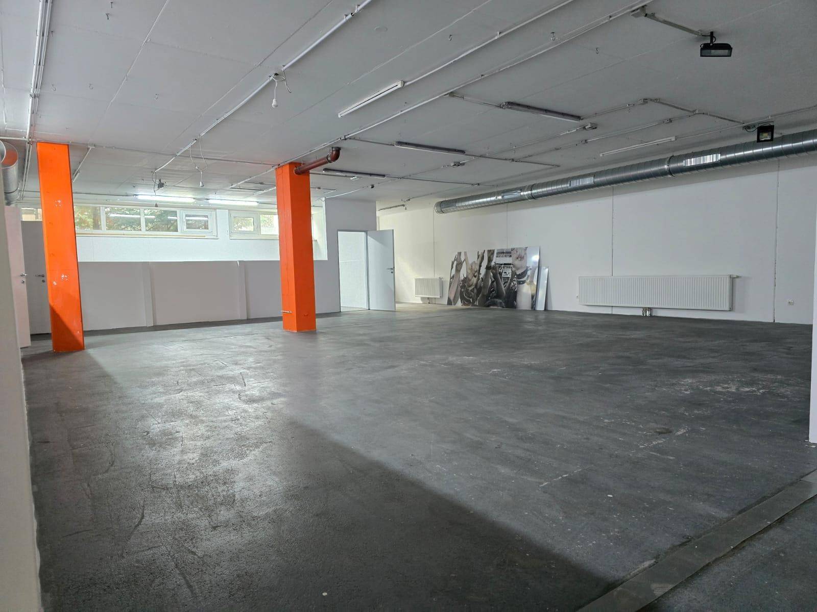 Großer, leerer Gewerberaum mit orangefarbenen Säulen, hohen Decken und Fenstern im oberen Wandbereich.