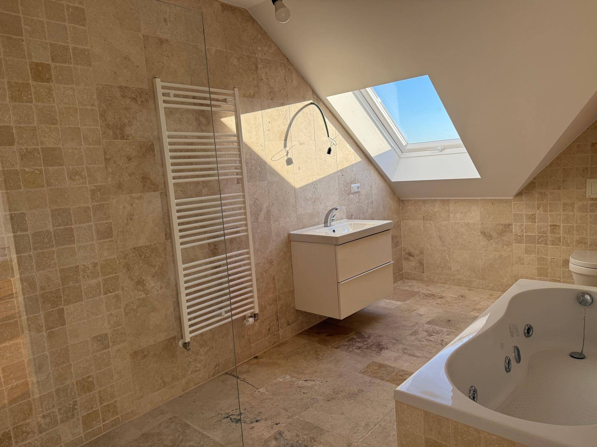 Helles Badezimmer mit Badewanne, beigen Fliesen und Dachfenster für viel Tageslicht.