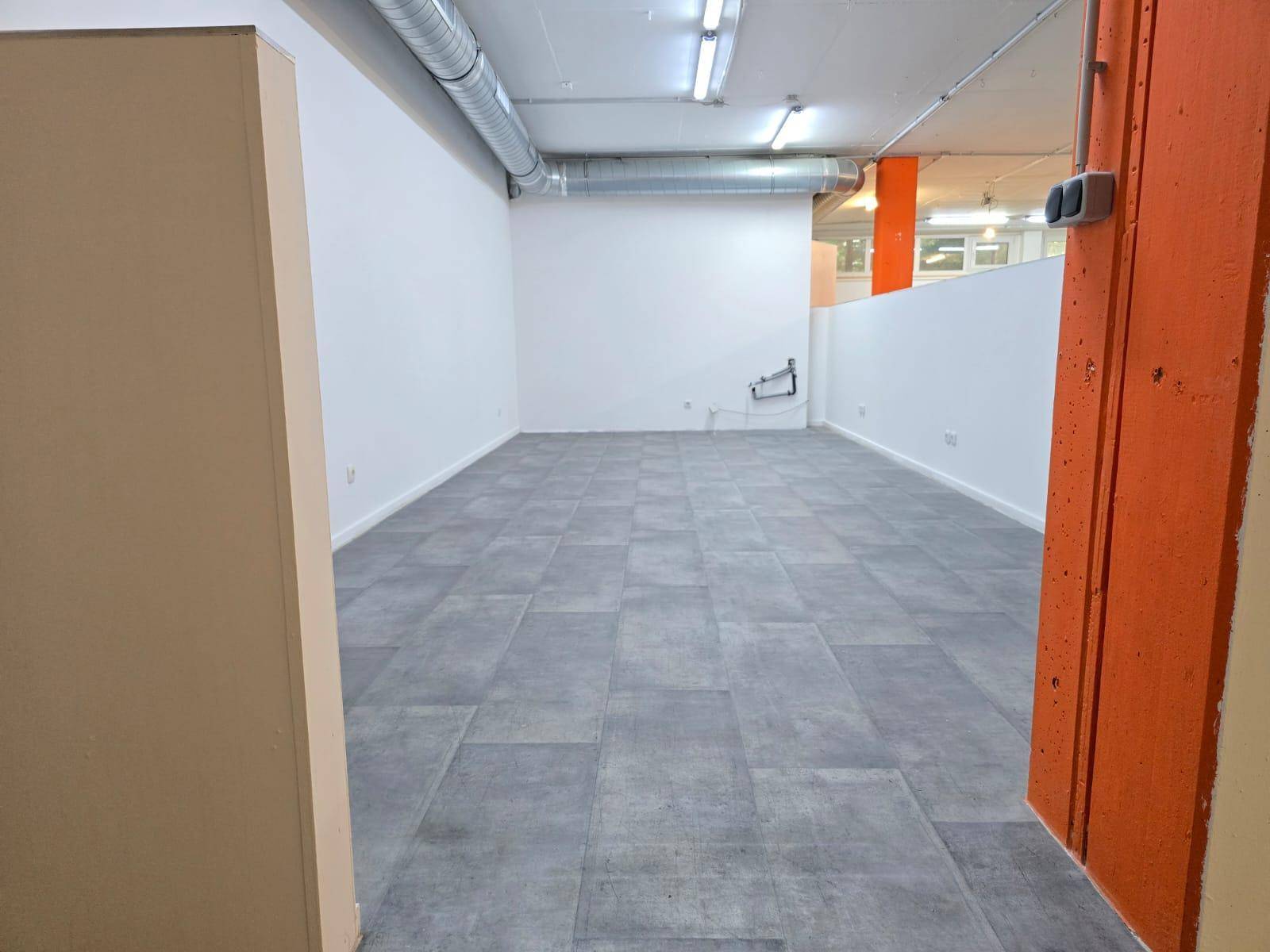 Heller Raum mit grauen Fliesen, weißen Wänden und sichtbaren Lüftungsrohren an der Decke.