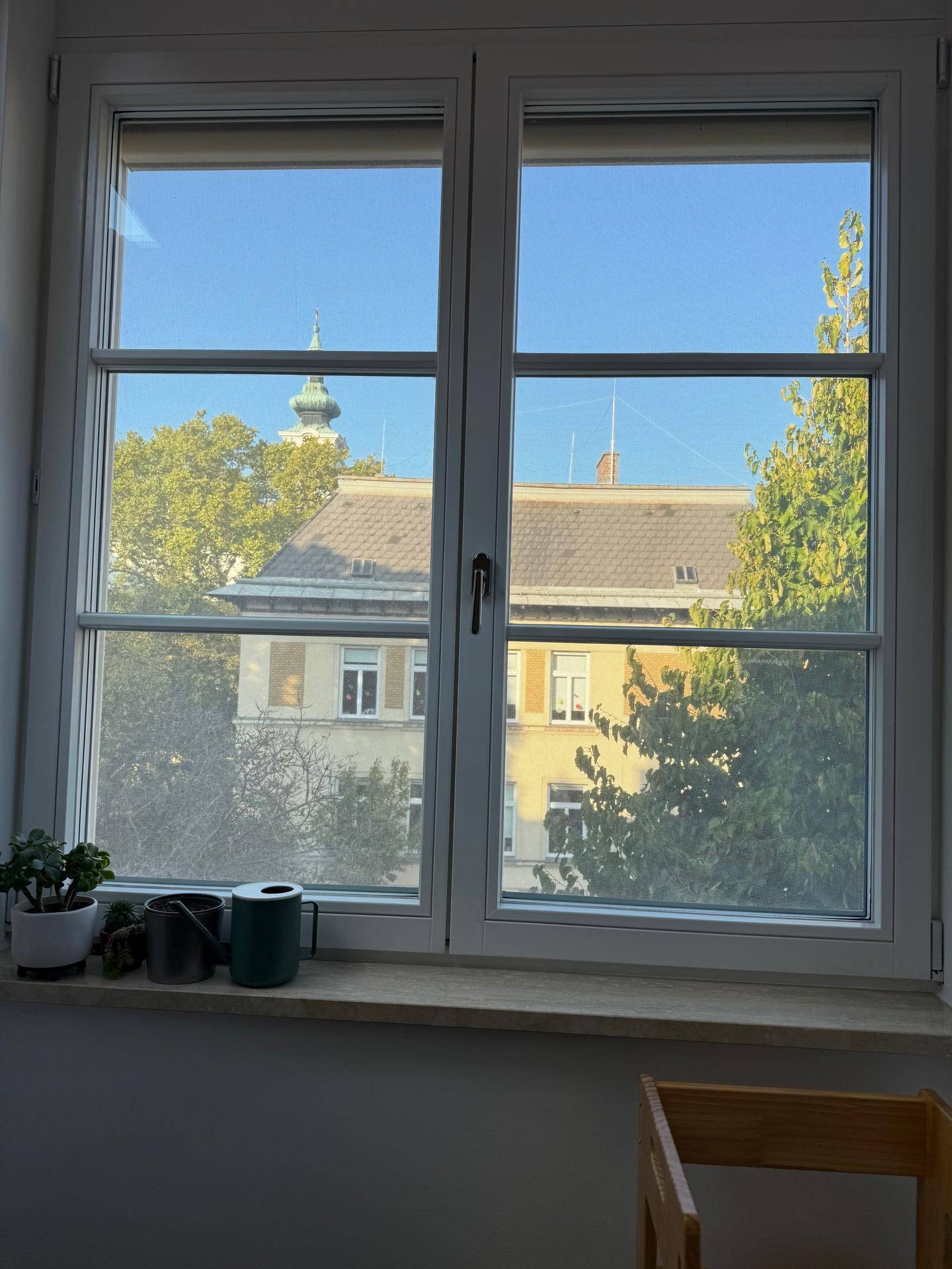 Fensterblick auf umliegende Gebäude, Bäume und einen Kirchturm in der Ferne.