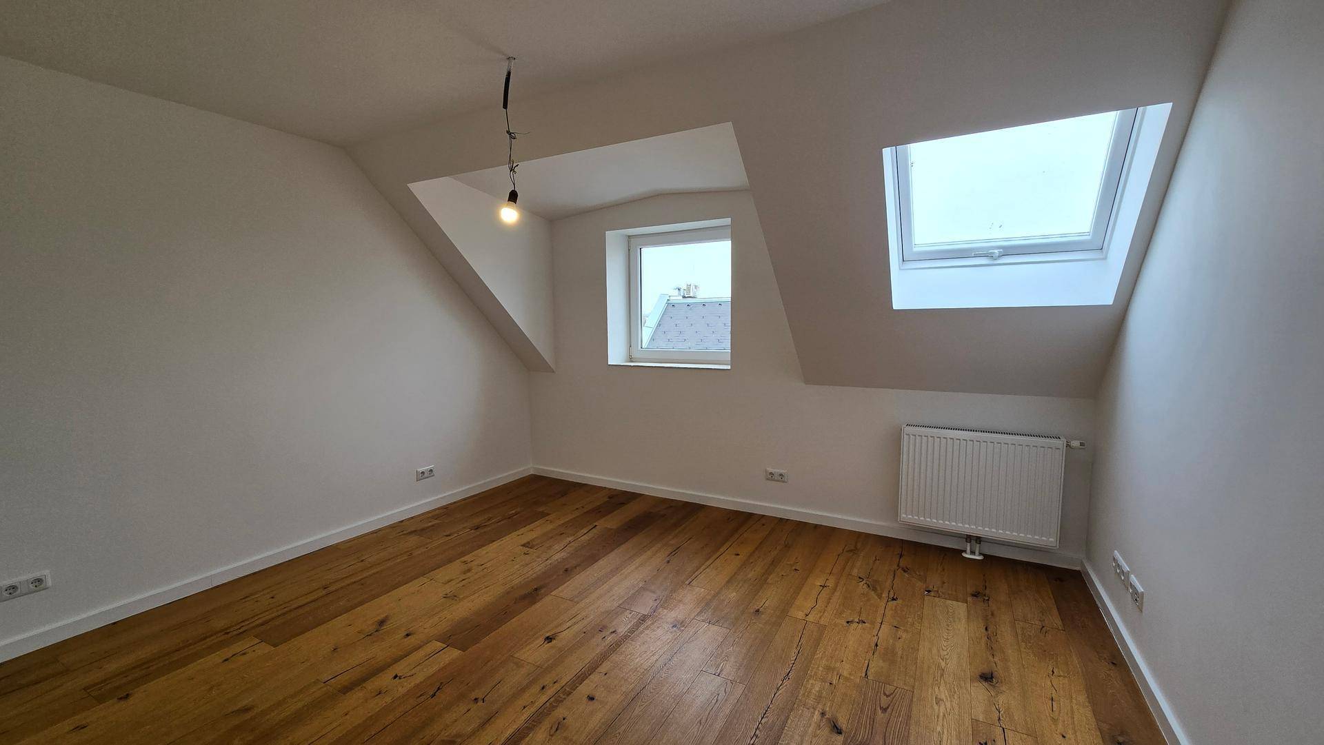 Schlafzimmer im Dachgeschoss mit Holzboden, Heizkörper und zwei Fenstern für Helligkeit.