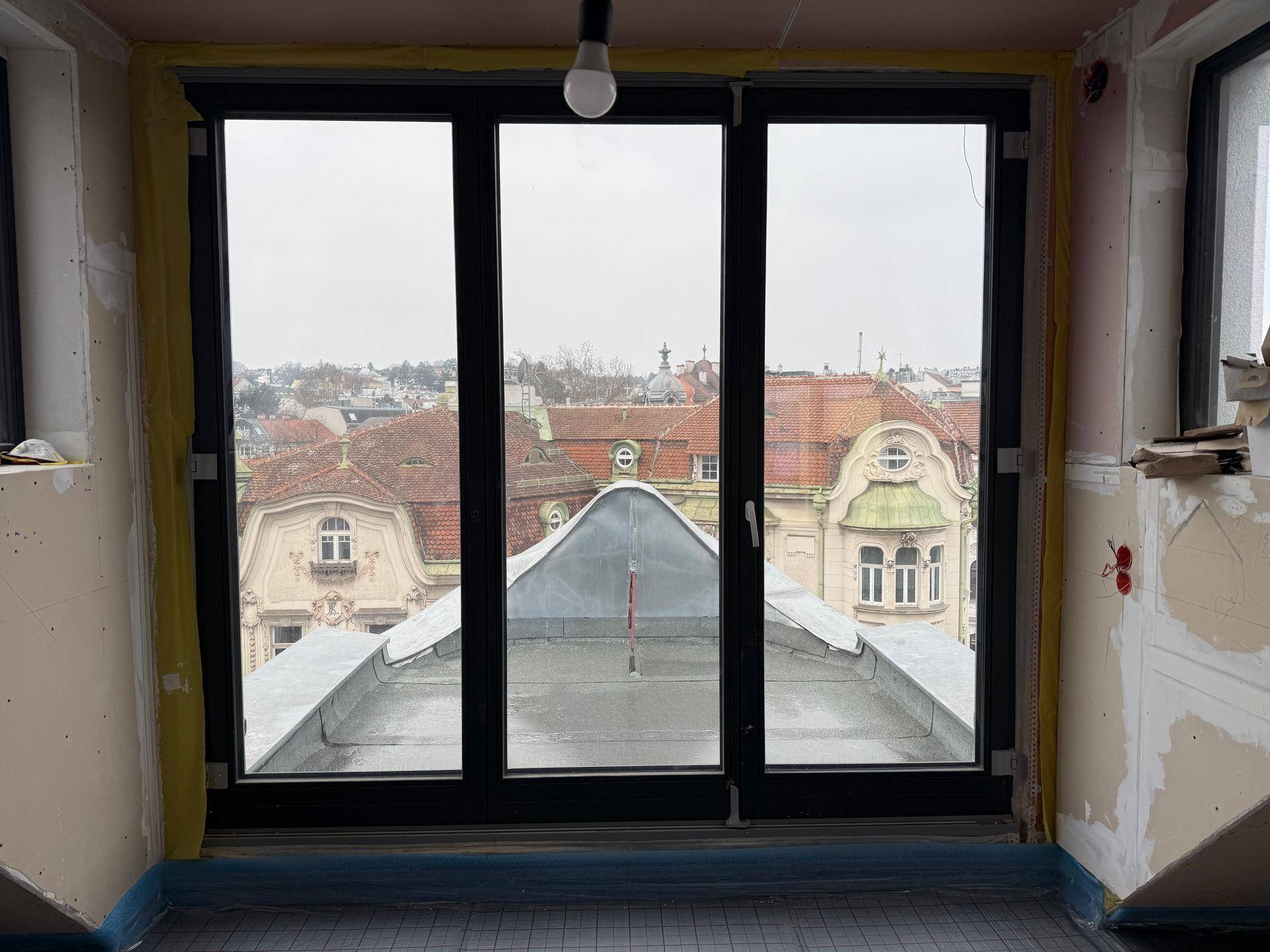 Innenraum im Bau mit großen Fenstern und Blick auf die Dächer der Stadt, Fußbodenheizung sichtbar.