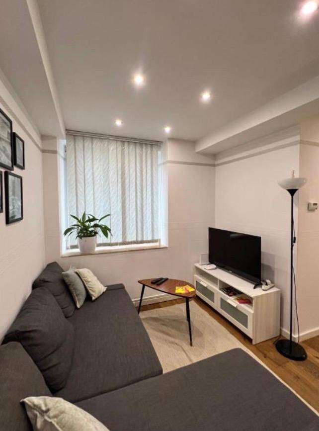Gemütlicher Wohnbereich mit grauem Ecksofa, Fernseher und Fenster mit vertikalen Jalousien.