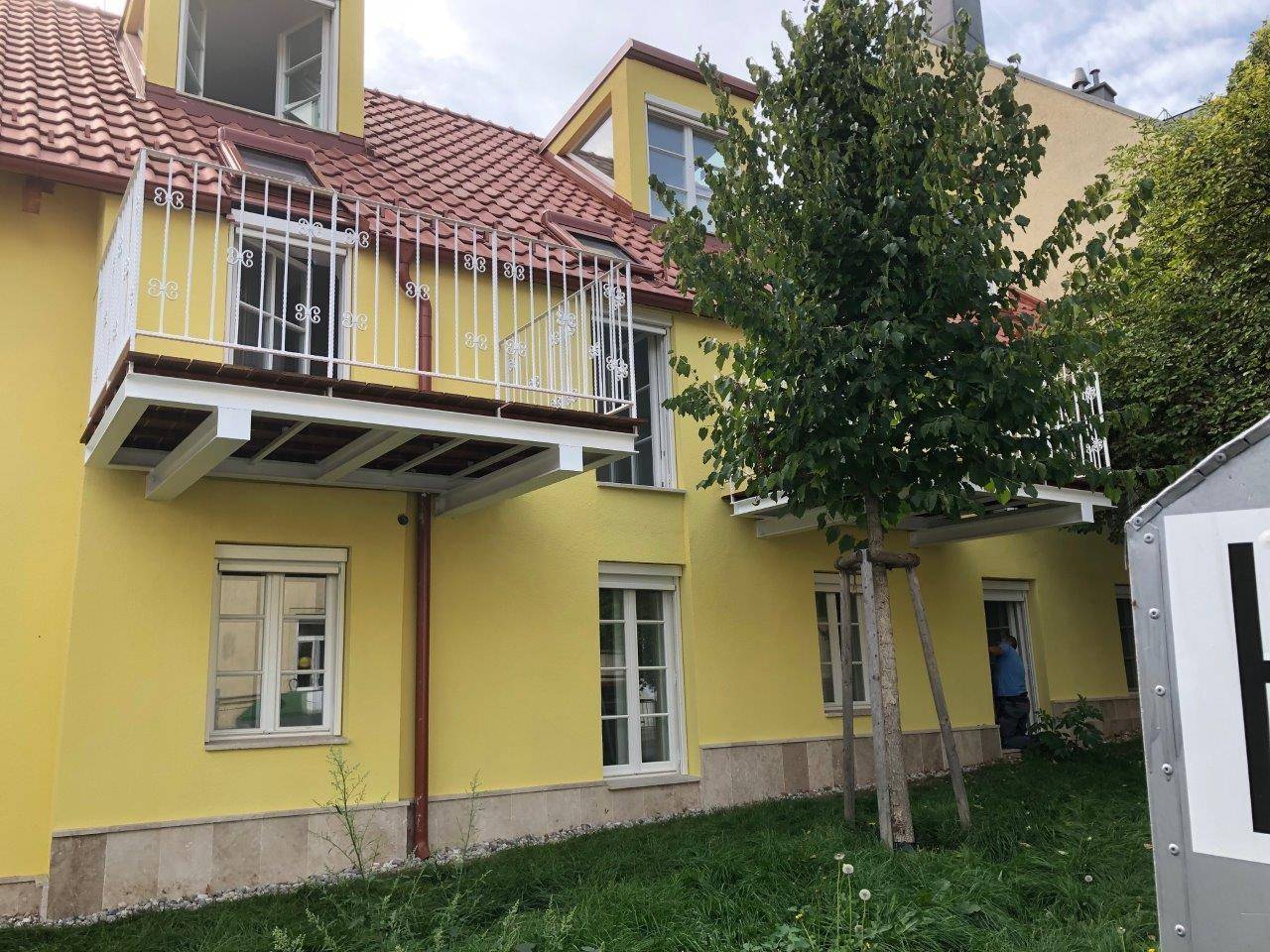 Gelbes Gebäude mit Balkon und rotem Ziegeldach, umgeben von Grünflächen und Bäumen.