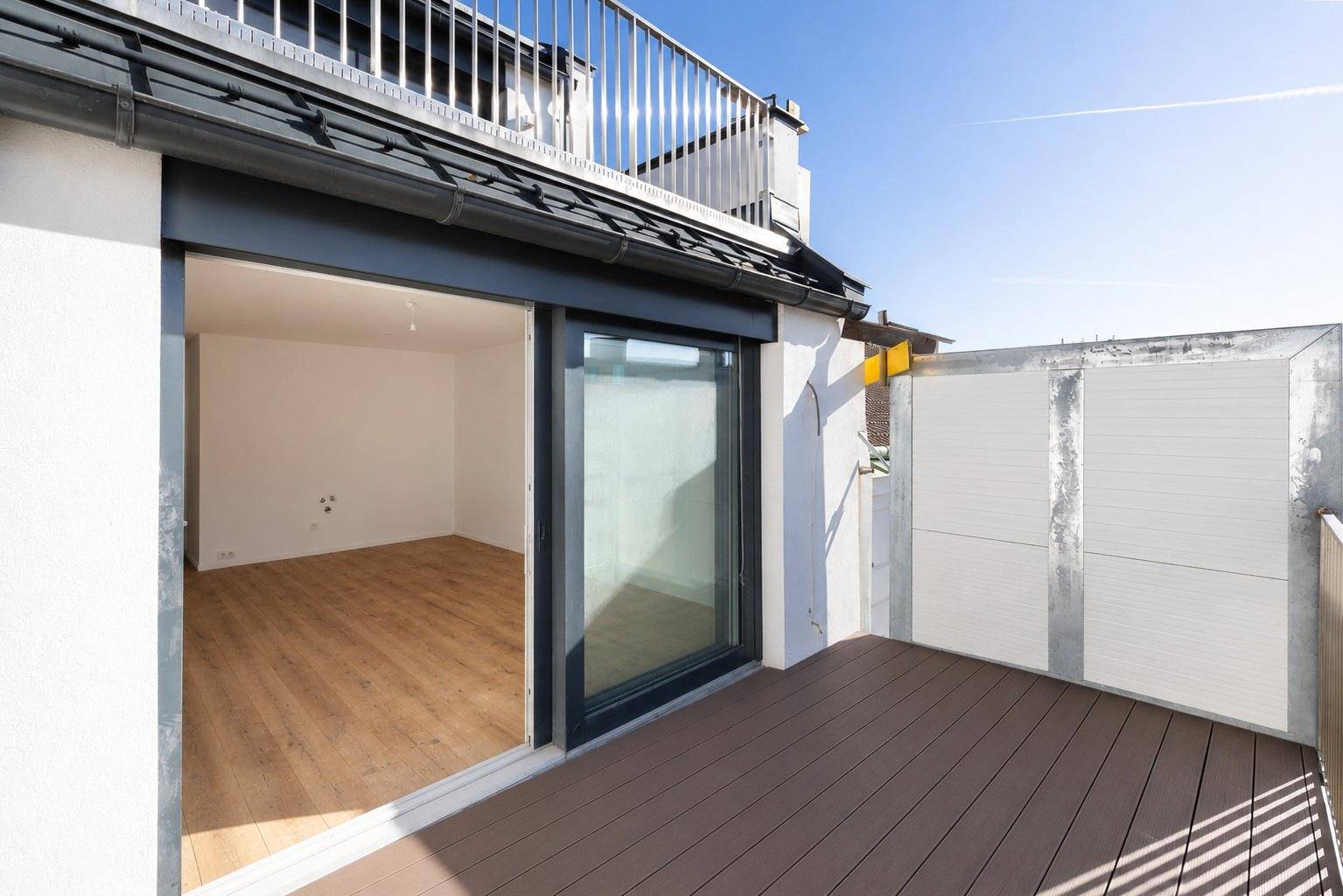 Geräumige Dachterrasse mit Holzboden und Zugang zu einem hellen, leeren Raum unter blauem Himmel.