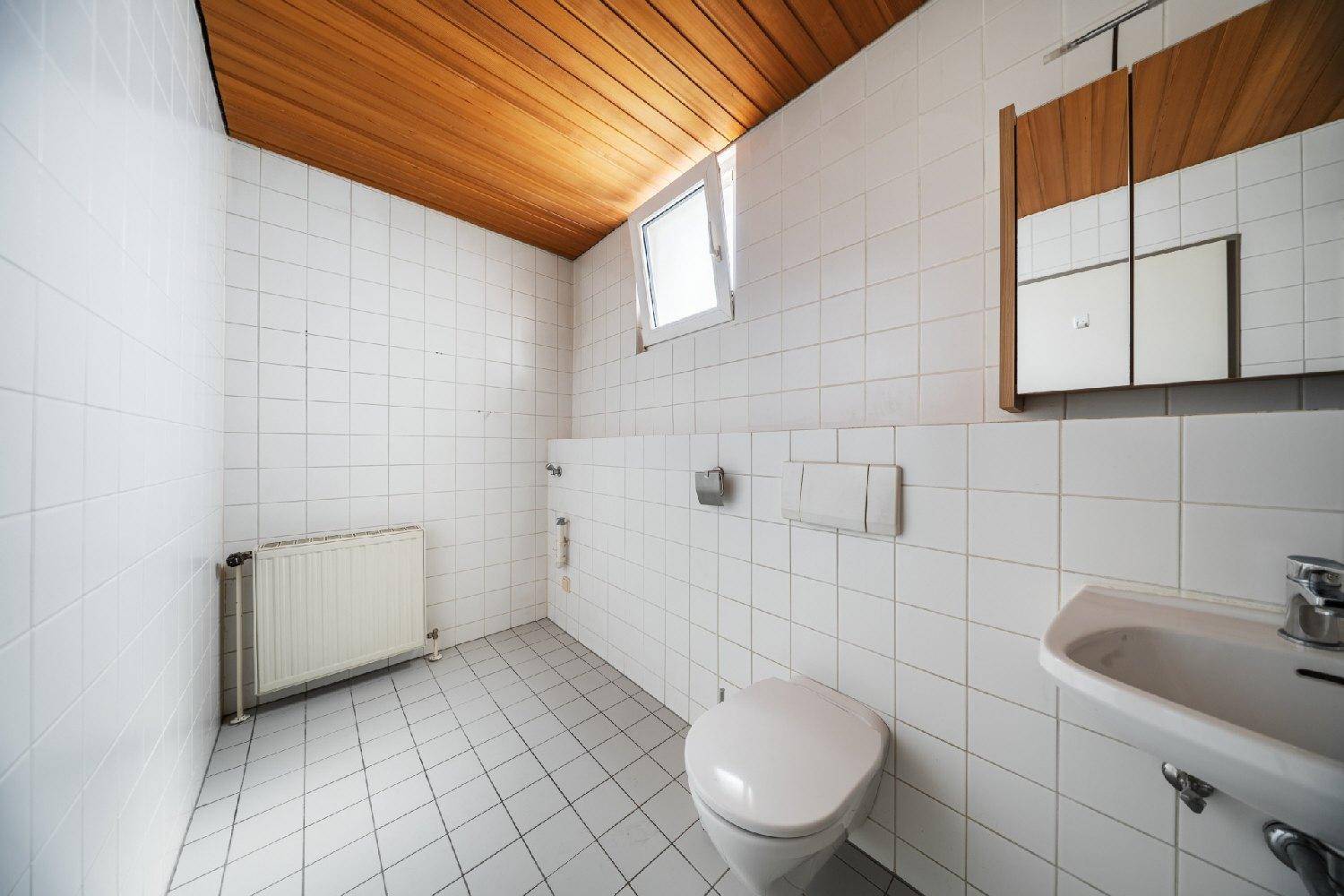 Einfaches WC mit weißen Fliesen, Holzdecke und kleinem Fenster für natürliche Belüftung.