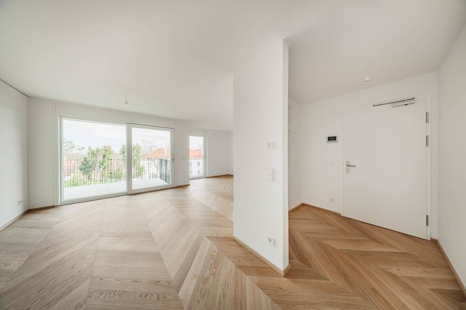Helles, leeres Wohnzimmer mit modernem Fischgrätparkett und Zugang zu einem Balkon.