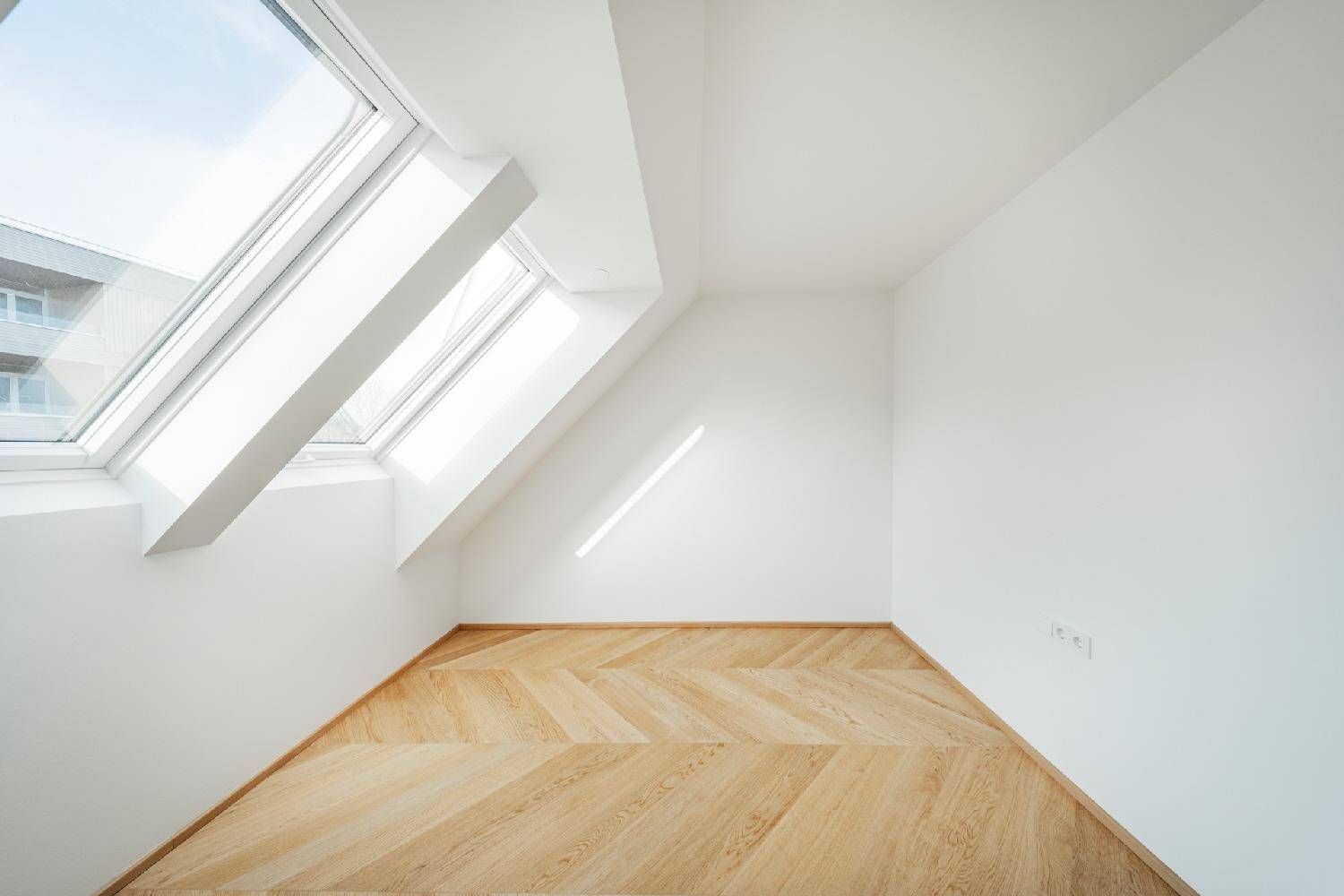 Leeres Zimmer im Dachgeschoss mit schrägen Wänden und zwei großen Dachfenstern für viel Tageslicht.