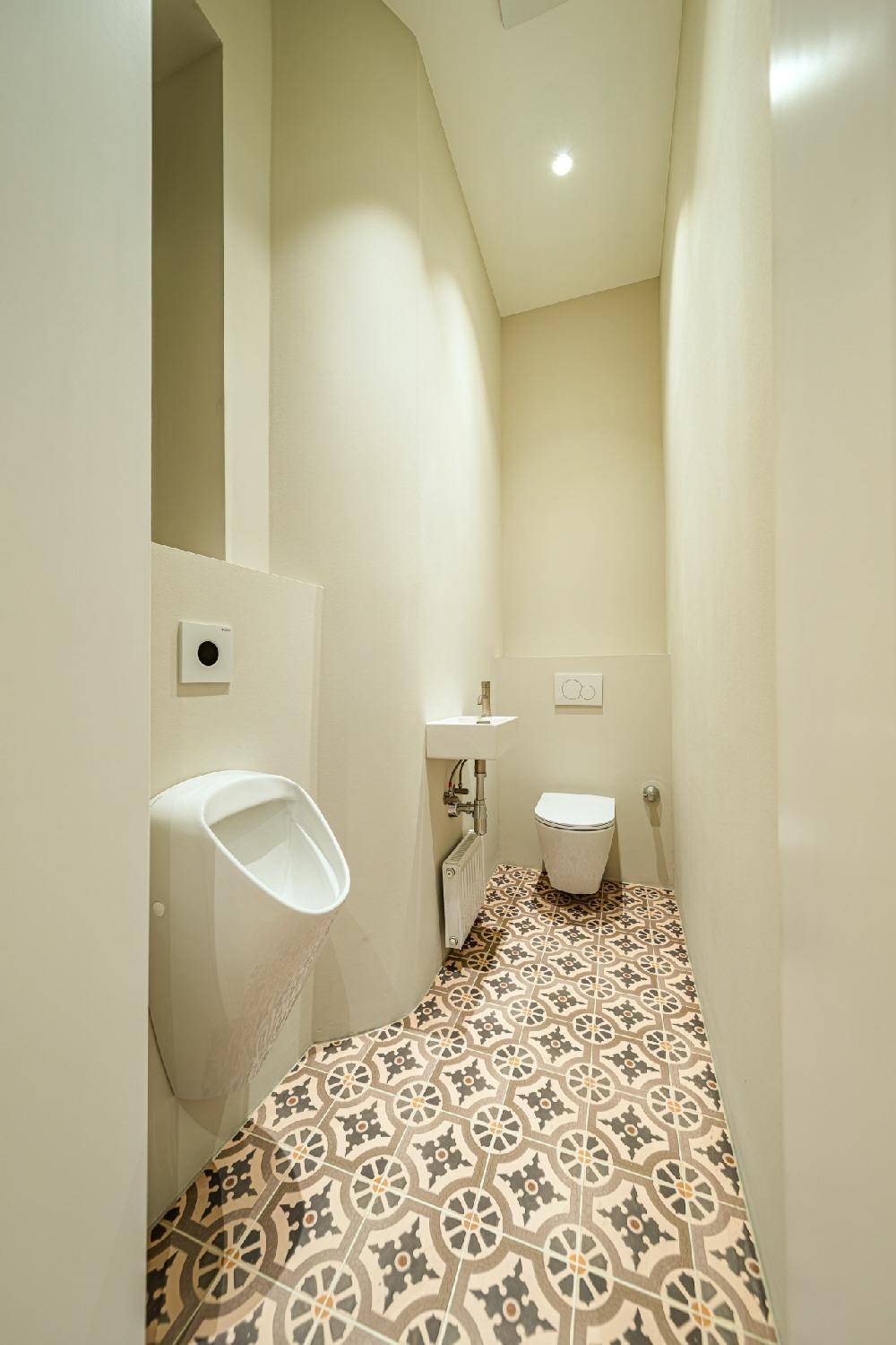 Modernes Gäste-WC mit Urinal, Wand-WC und gemusterten Fliesenboden in einem schmalen Raum.