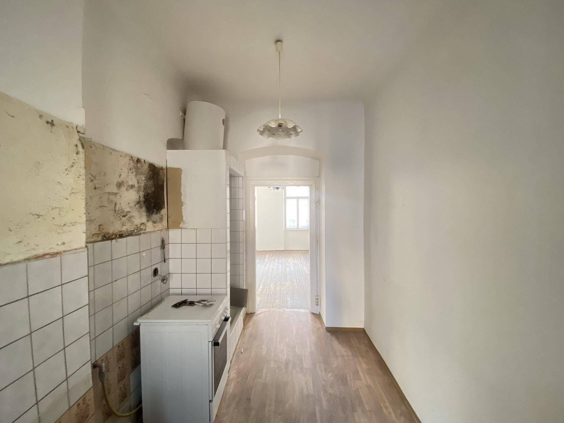 Schmale Küche mit einfacher Ausstattung, sichtbaren Fliesenresten und Durchgang zu einem weiteren Zimmer.