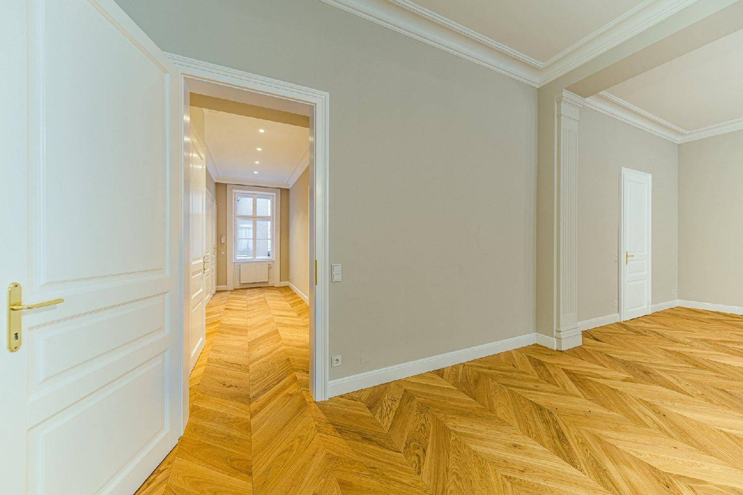 Heller Flur mit Fischgrätparkett und weißen Türen, der zu einem Zimmer mit Fenster und weiteren Räumen führt.