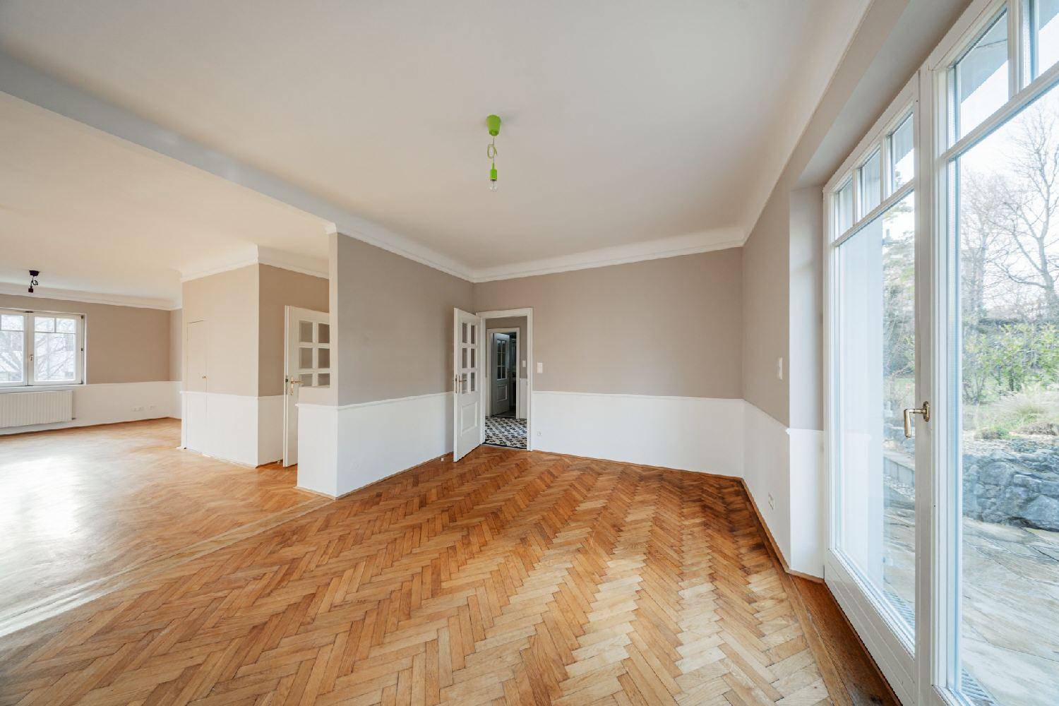 Großzügiges Wohnzimmer mit Fischgrätparkett, hohen Fenstern und offenem Blick in den angrenzenden Flur.