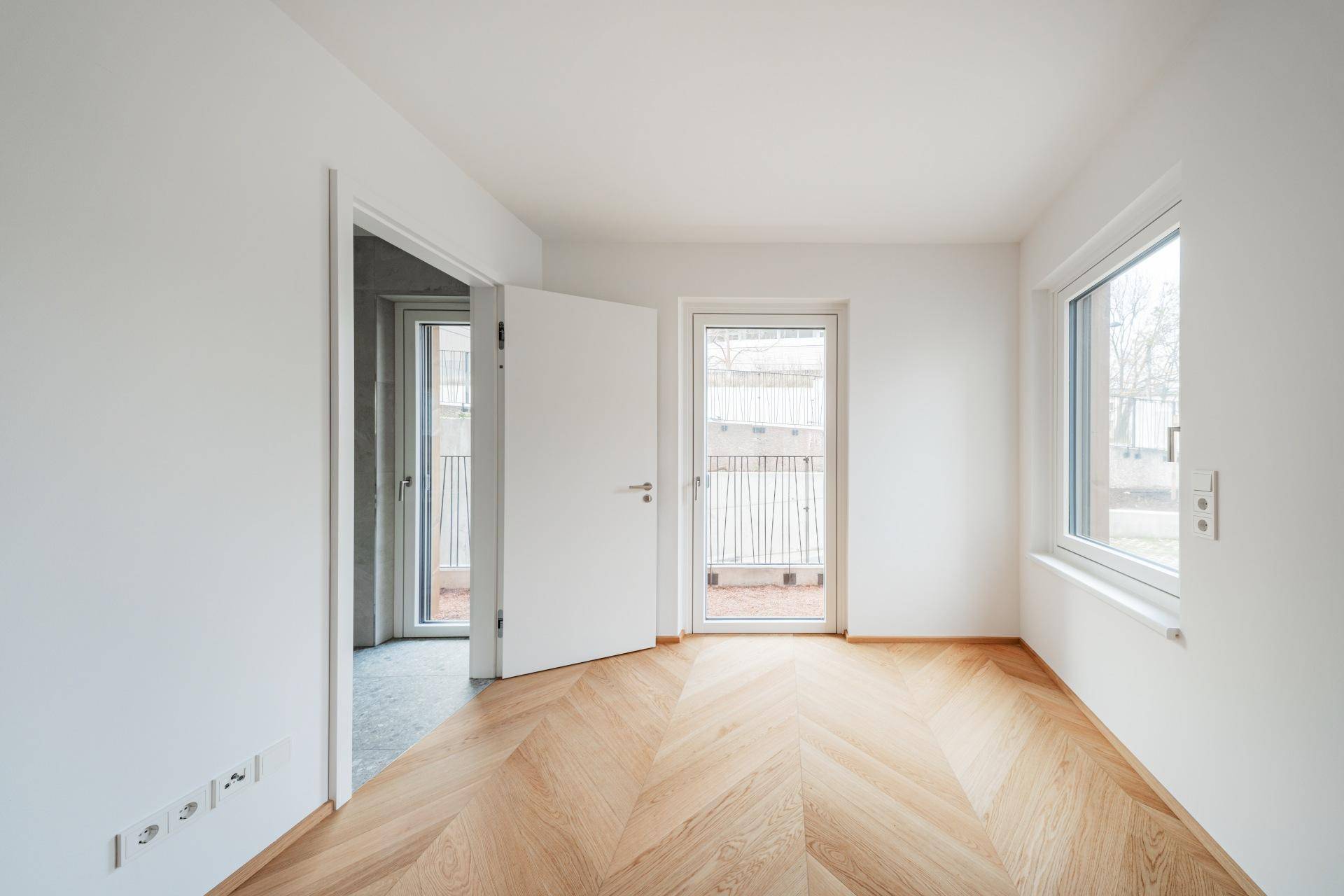 Helles Schlafzimmer mit Fischgrätparkett, Fenster und Zugang zu einem Außenbereich.