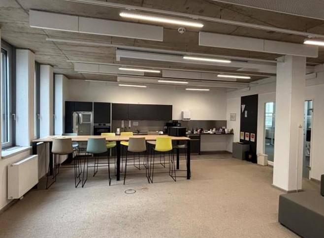 Moderne Büroküche mit langer Theke, bunten Hockern, dunklen Schränken und freiliegender Betondecke.