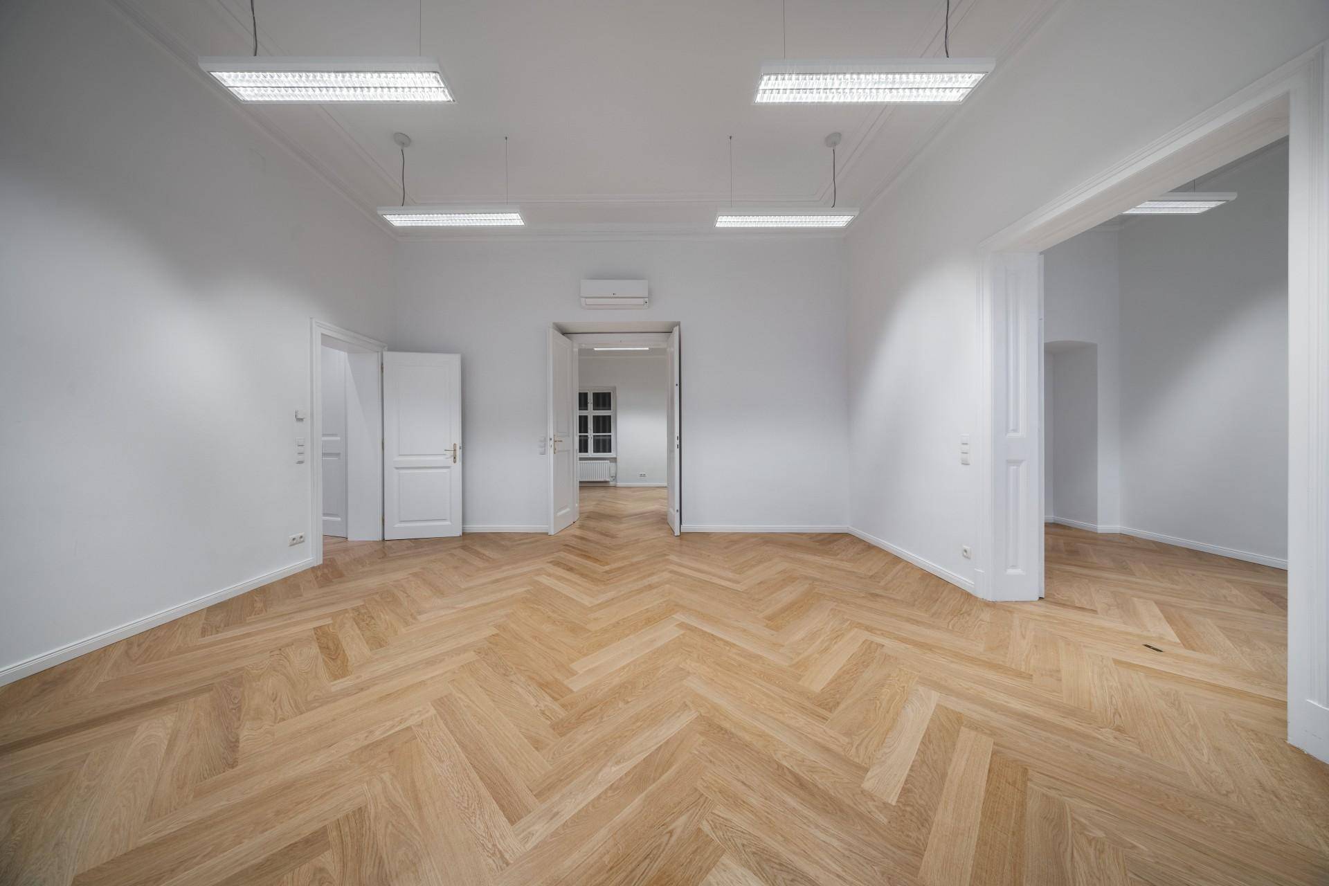 Großer, heller Raum mit Fischgrätparkett, weißen Wänden und mehreren Türen.