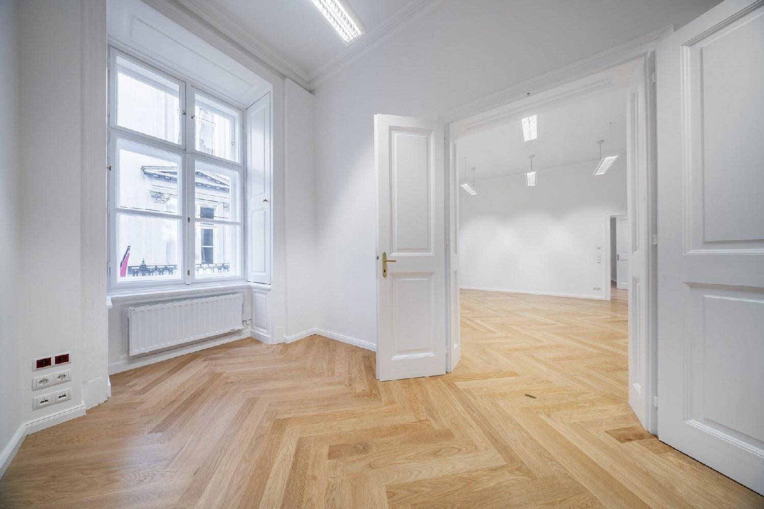 Heller, leerer Raum mit Fischgrätparkett, großem Fenster und Heizkörper unter dem Fenster.
