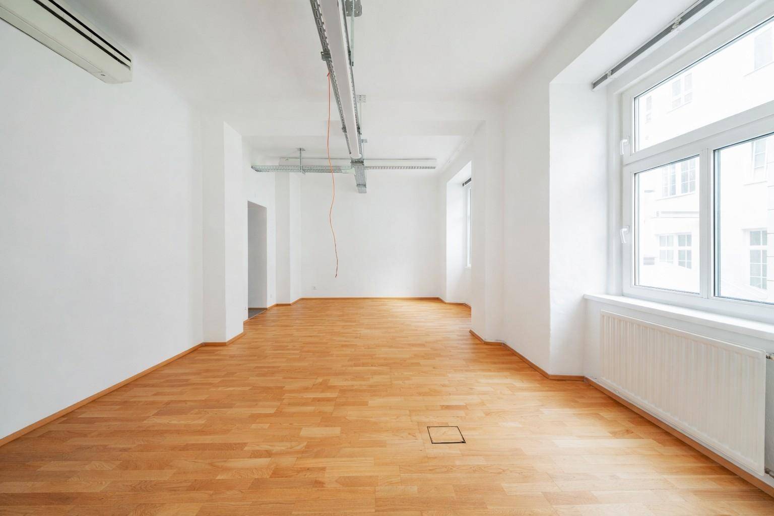 Langer, heller Büroraum mit Holzboden, Klimaanlage und großen Fenstern, die viel Tageslicht hereinlassen.