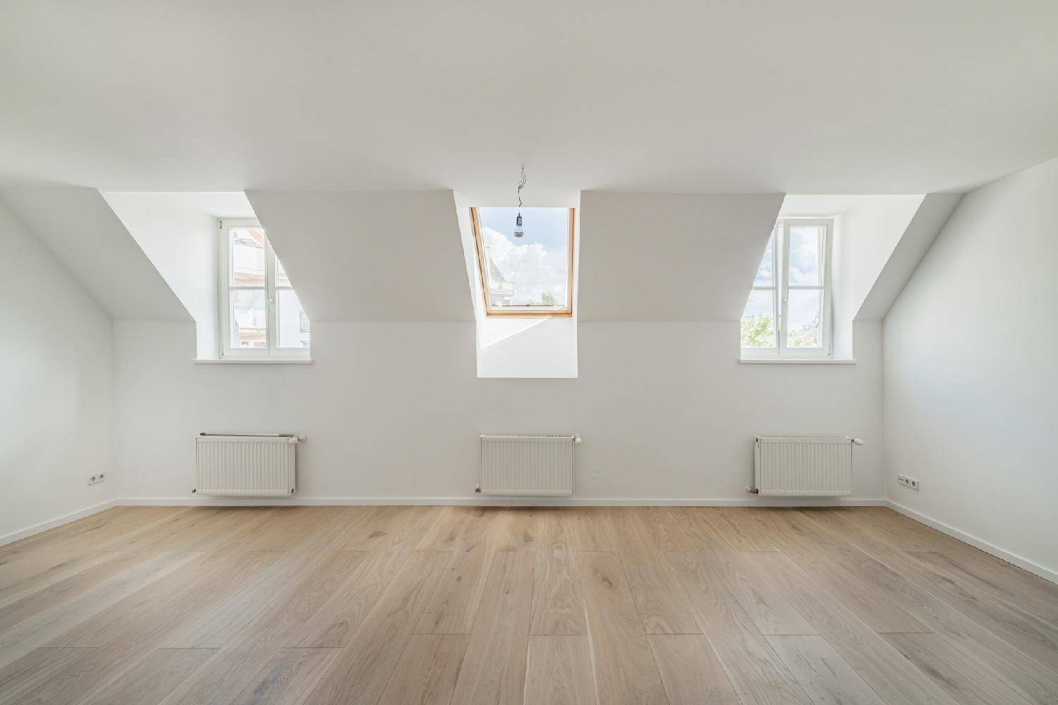 Heller, leerer Dachbodenraum mit schrägen Wänden, Holzboden und mehreren Fenstern, inklusive Dachfenster.
