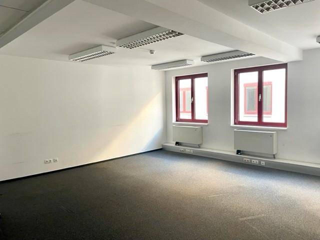 Heller, leerer Büroraum mit Teppichboden, weißen Wänden, roten Fensterrahmen und integrierter Deckenbeleuchtung.