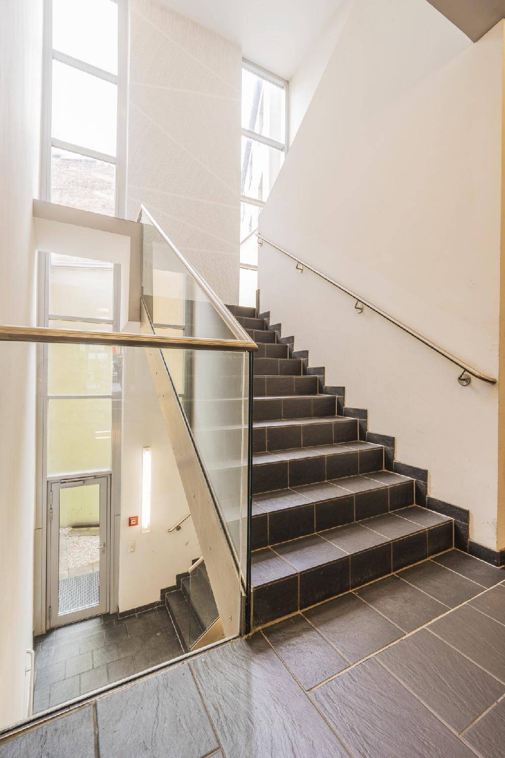Modernes Treppenhaus mit Fliesenstufen, Glasgeländer und großen Fenstern für viel Tageslicht.