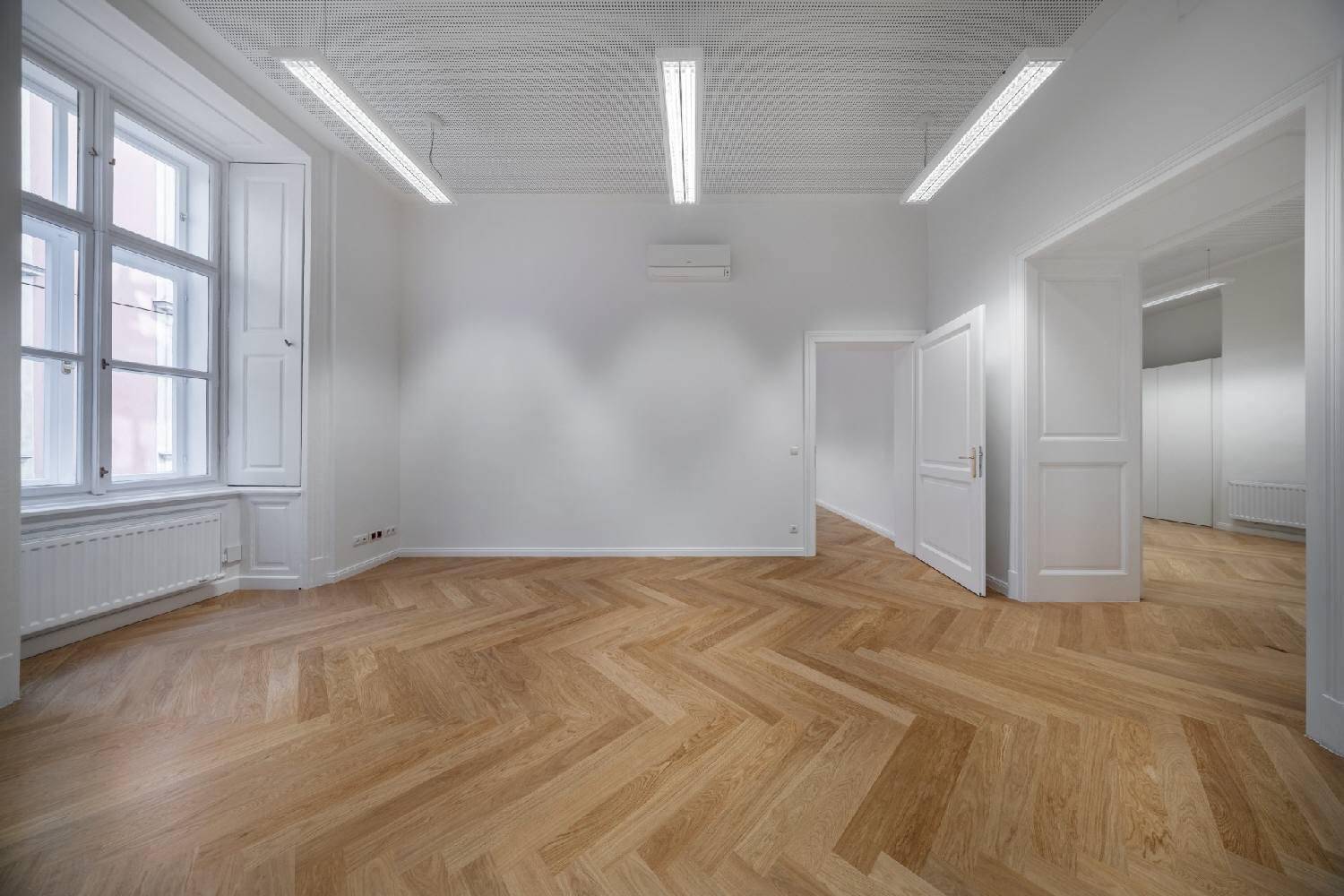 Heller, leerer Raum mit Fischgrätparkett, hohen Fenstern mit Fensterläden und Klimaanlage.