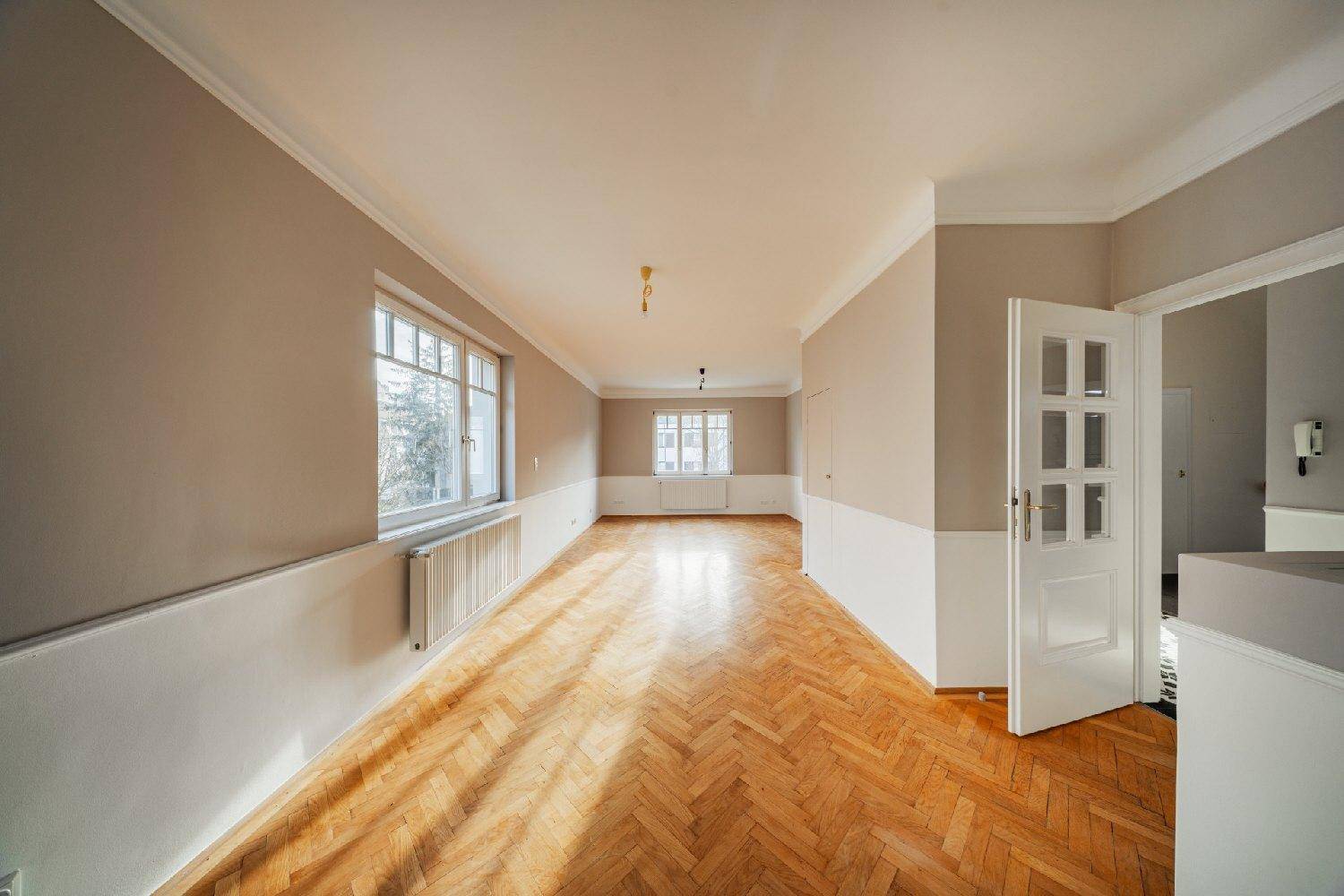 Geräumiges Wohnzimmer mit Fischgrätparkett, hohen Decken und viel Tageslicht durch große Fenster.