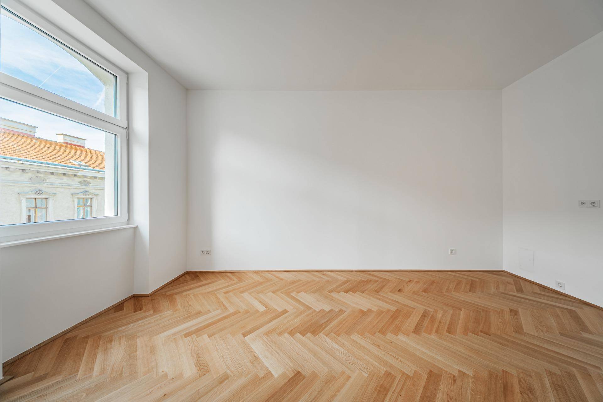 Lichtdurchfluteter Raum mit großem Fenster und klassischem Fischgrätparkett, ideal für Wohn- oder Schlafzimmer.