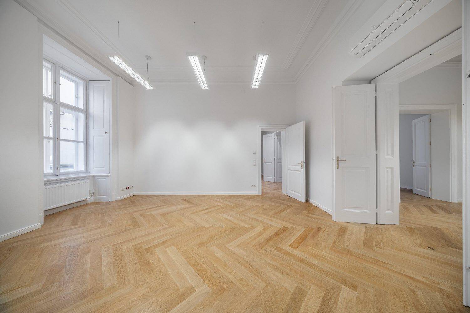 Geräumiger, heller Raum mit Fischgrätparkett, hohen Fenstern mit Fensterläden und modernen Deckenleuchten.
