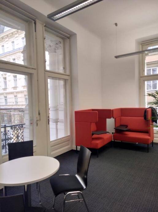 Gemütlicher Loungebereich mit roten Sesseln, einem kleinen Tisch und großen Fenstern mit Blick auf die Stadt.
