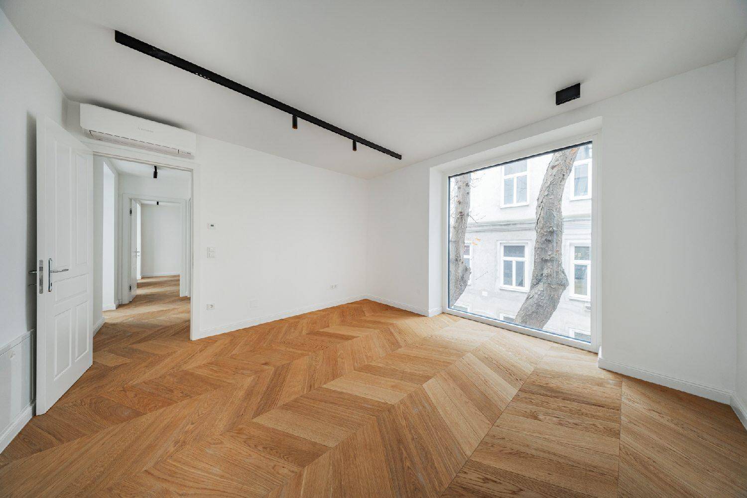 Helles Wohnzimmer mit Fischgrätparkett, großer Fensterfront und Klimaanlage.