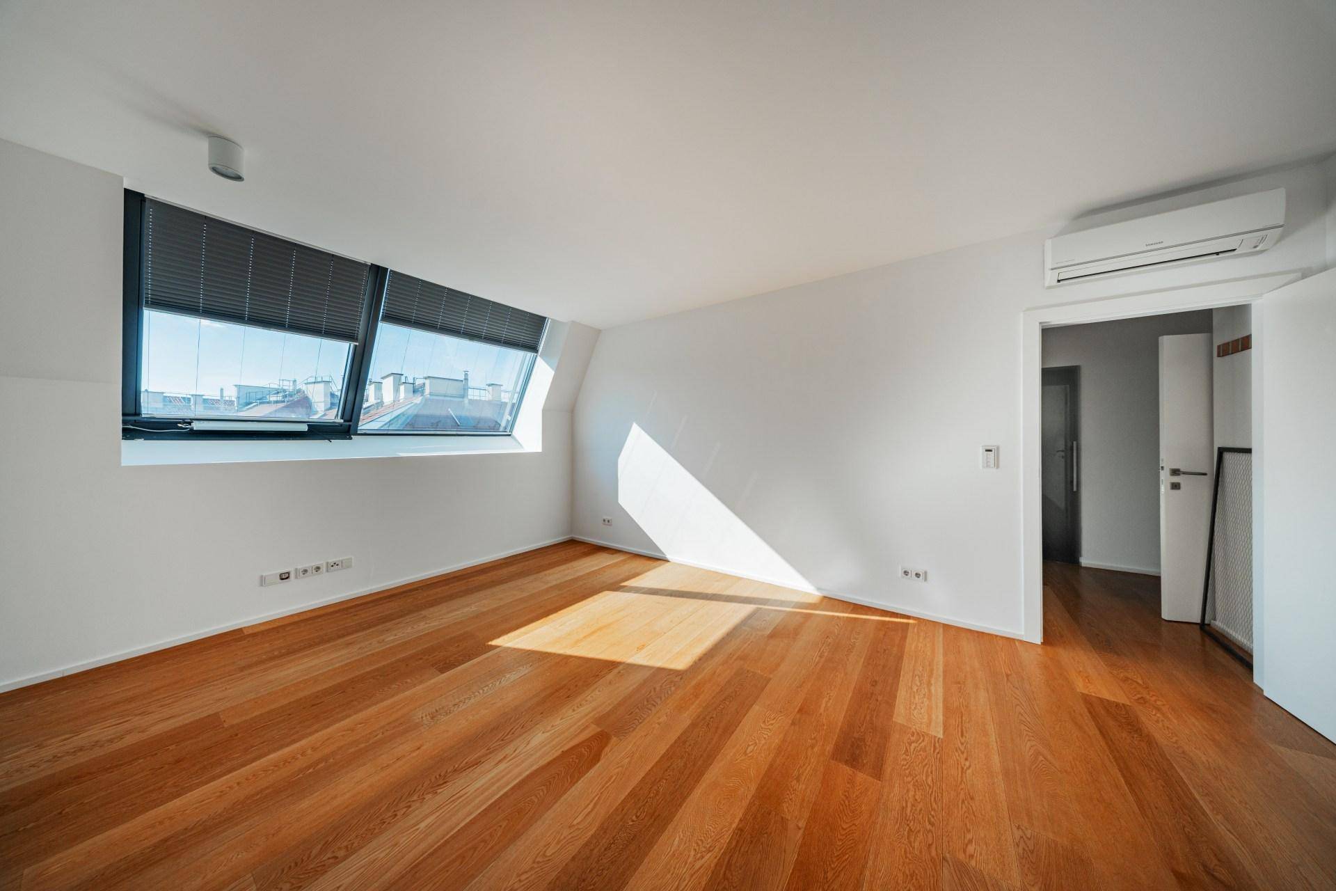 Helles Schlafzimmer mit Holzboden, schrägen Wänden und einem Fenster mit Blick auf die Umgebung.