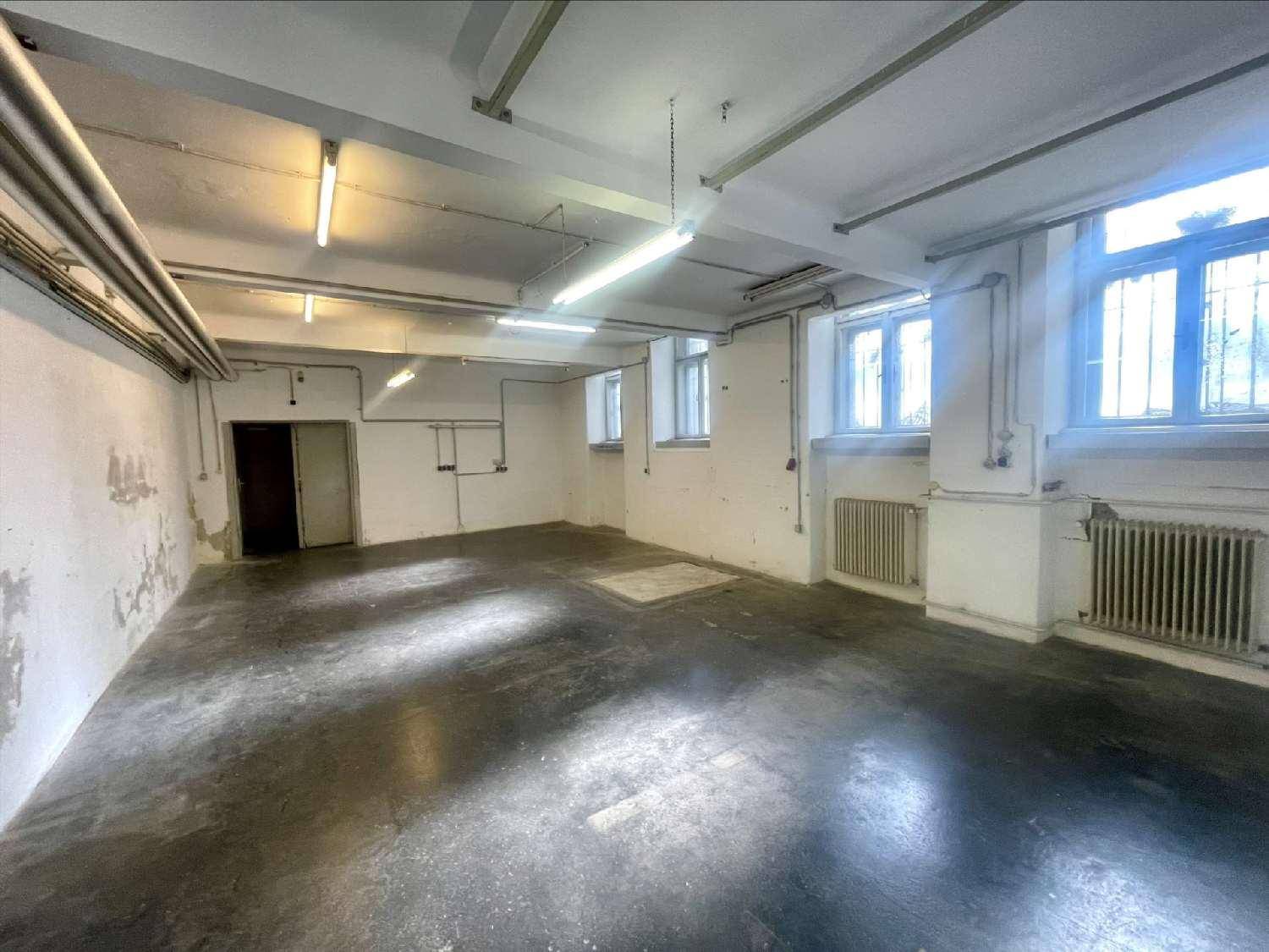 Großer, leerer Lagerraum mit hohen Decken, sichtbaren Rohren und Fenstern, die Tageslicht hereinlassen.