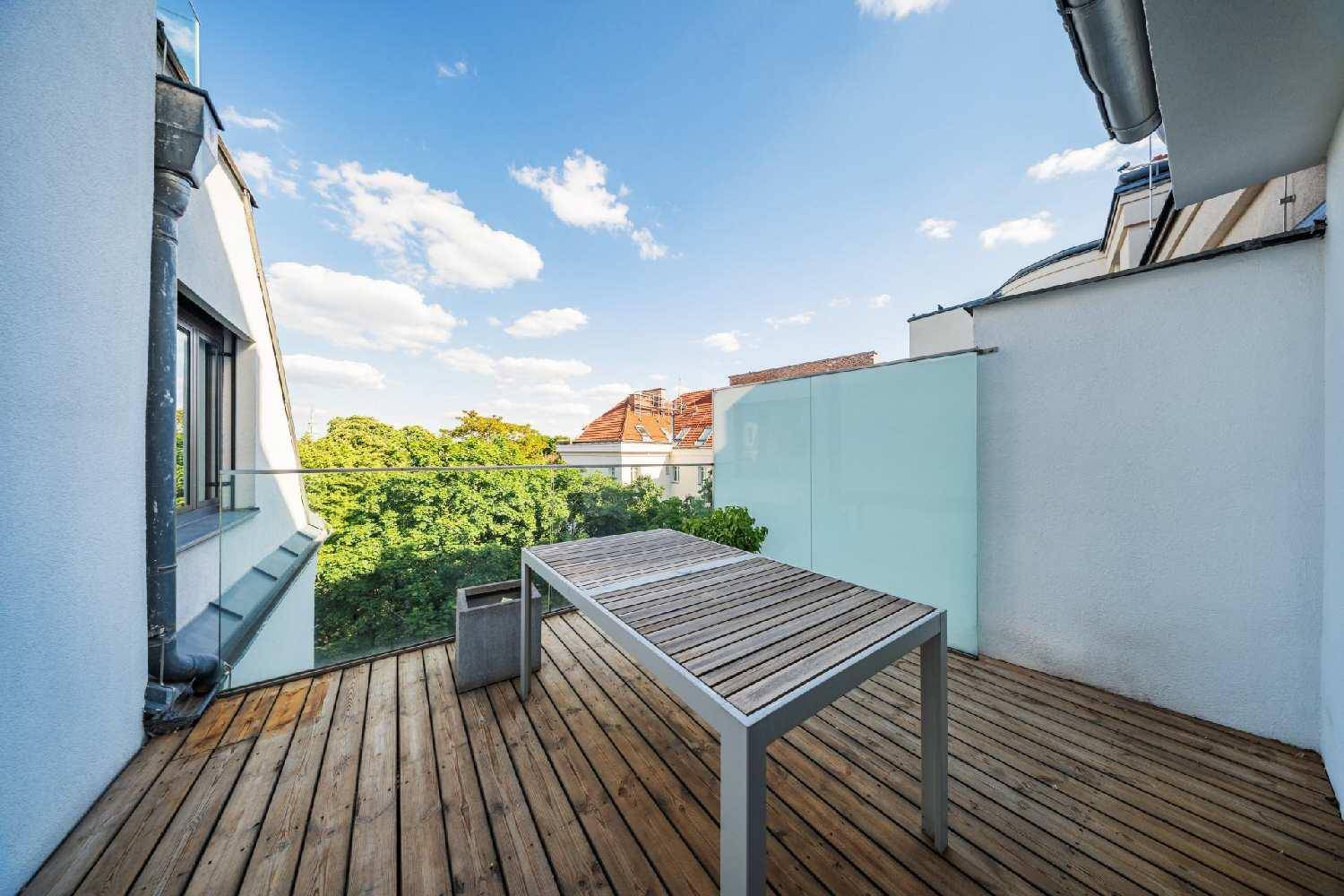 Sonniger Terrassenbereich mit Holzboden und Esstisch, umgeben von einem Glasgeländer und Bäumen.