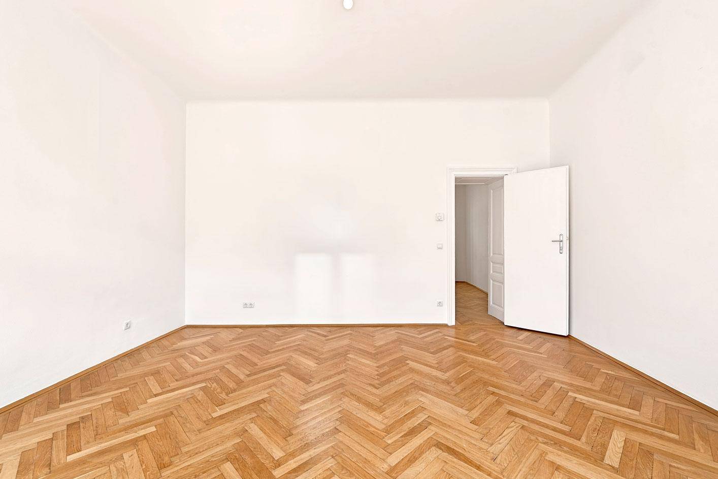 Leeres Zimmer mit Fischgrätparkett und einer offenen Tür, die den Blick in einen weiteren Raum freigibt.