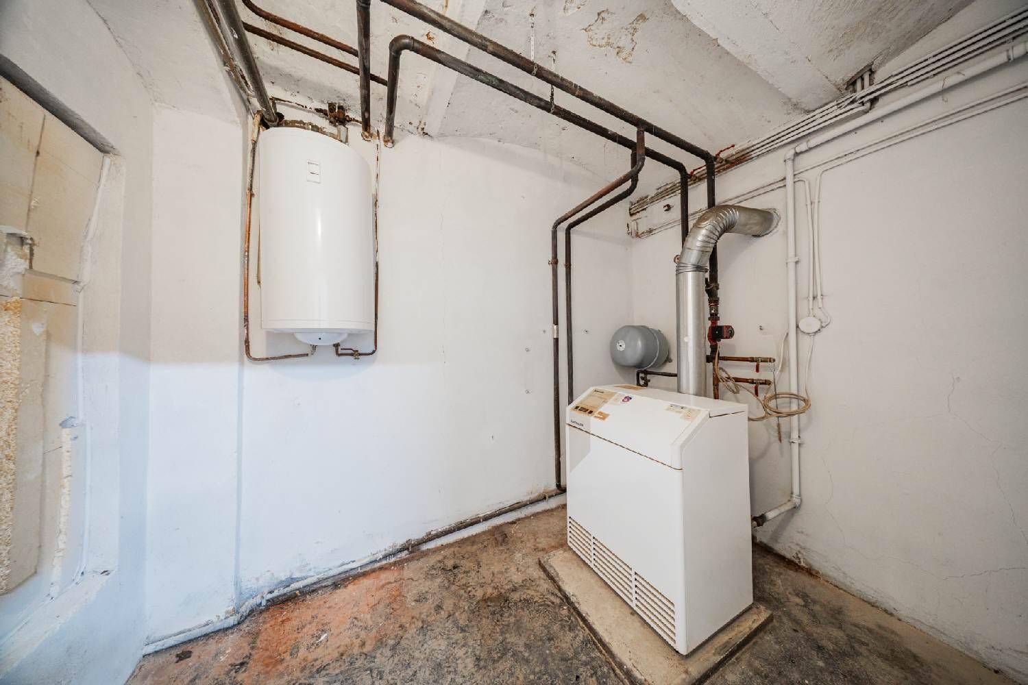 Technikraum mit Heizungsanlage, Warmwasserspeicher und sichtbaren Rohren an den Wänden.