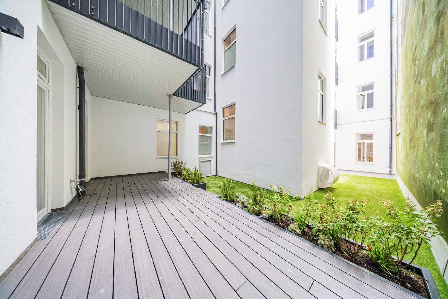 Geräumige Terrasse mit grauen Dielen, Pflanzenbeeten und Blick auf die moderne Gebäudefassade.