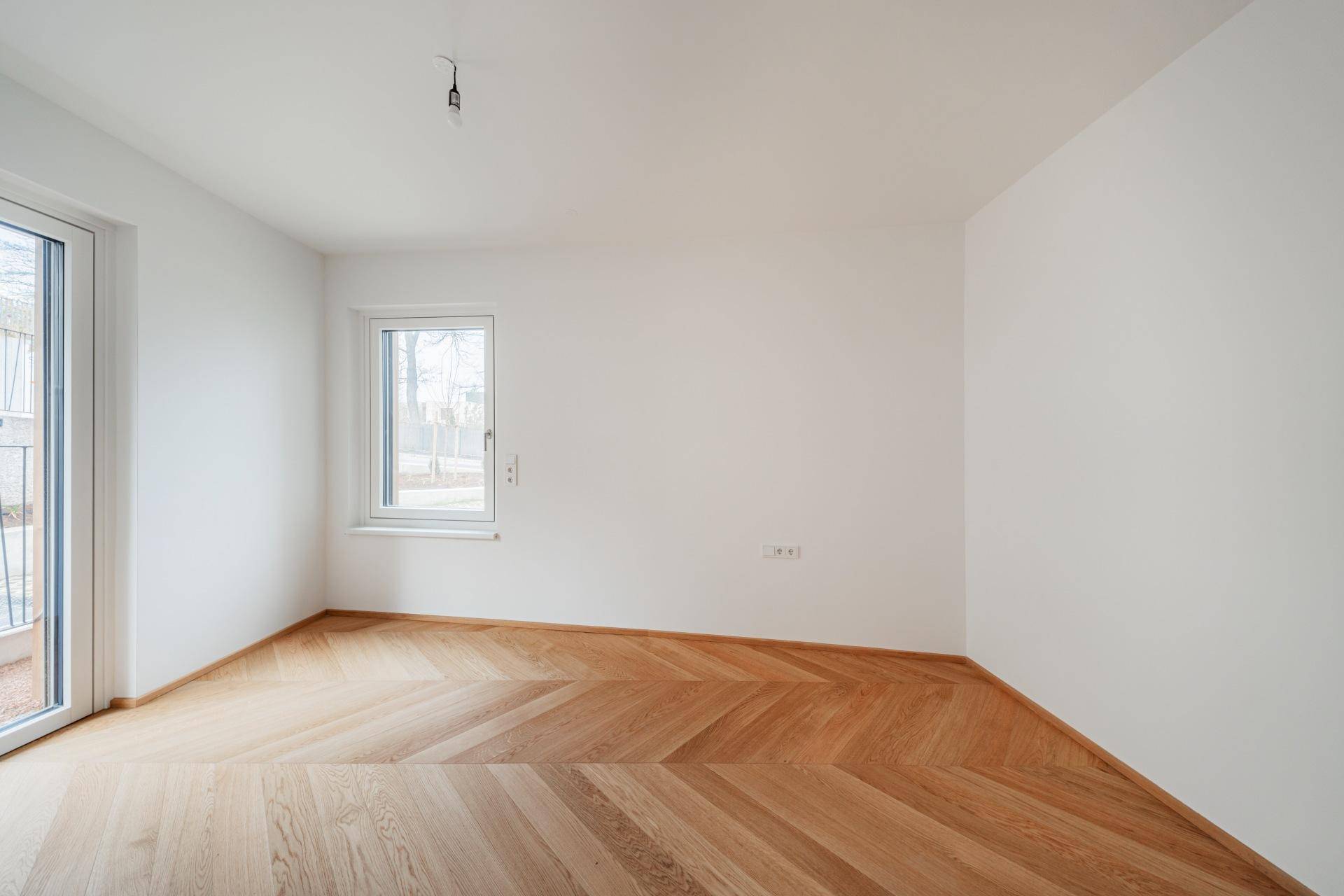 Leeres Zimmer mit hellem Fischgrätparkett und einem großen Fenster für viel Tageslicht.