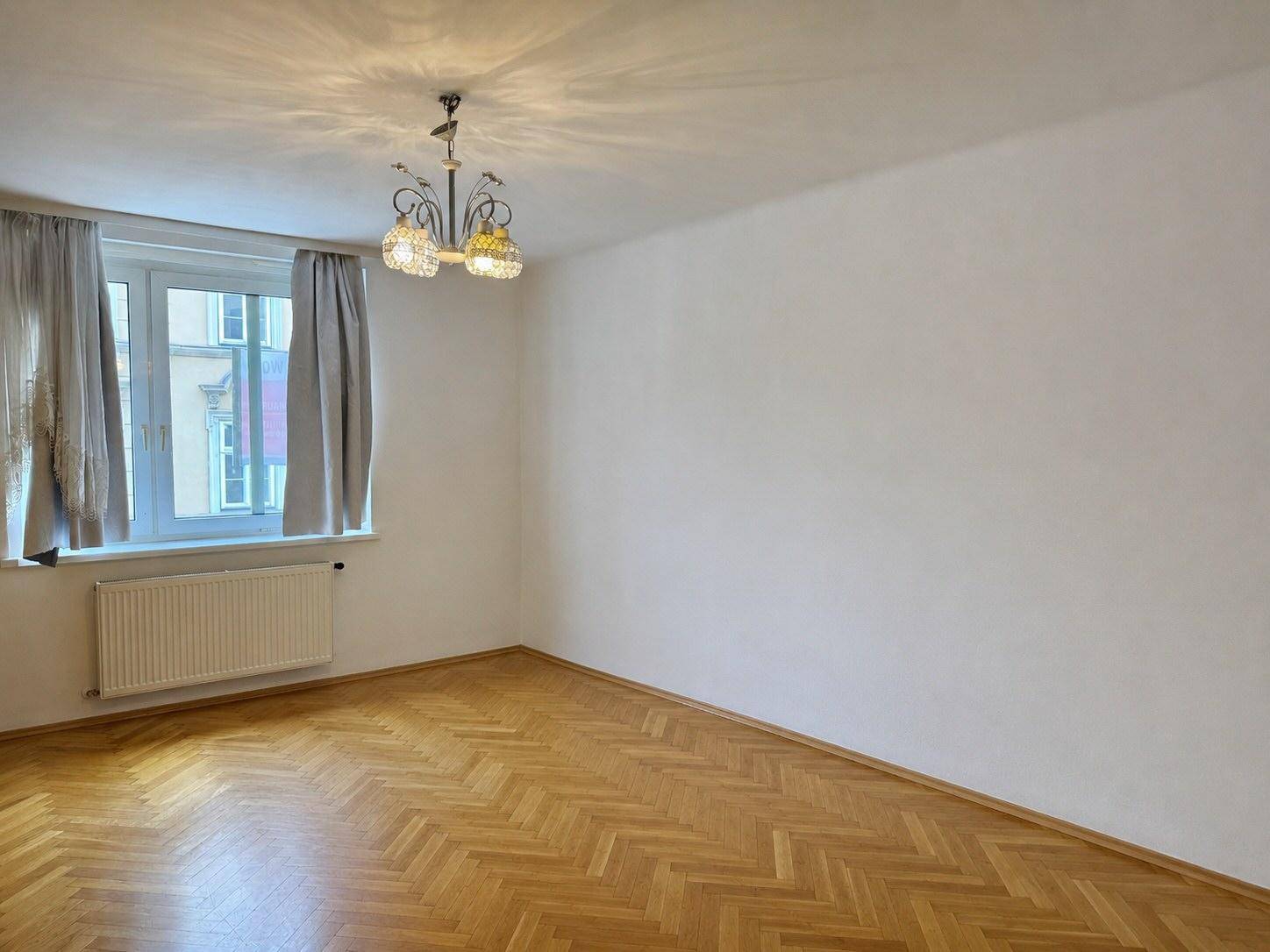Helles Zimmer mit Fischgrätparkett, großem Fenster und Heizkörper unter dem Fenster.