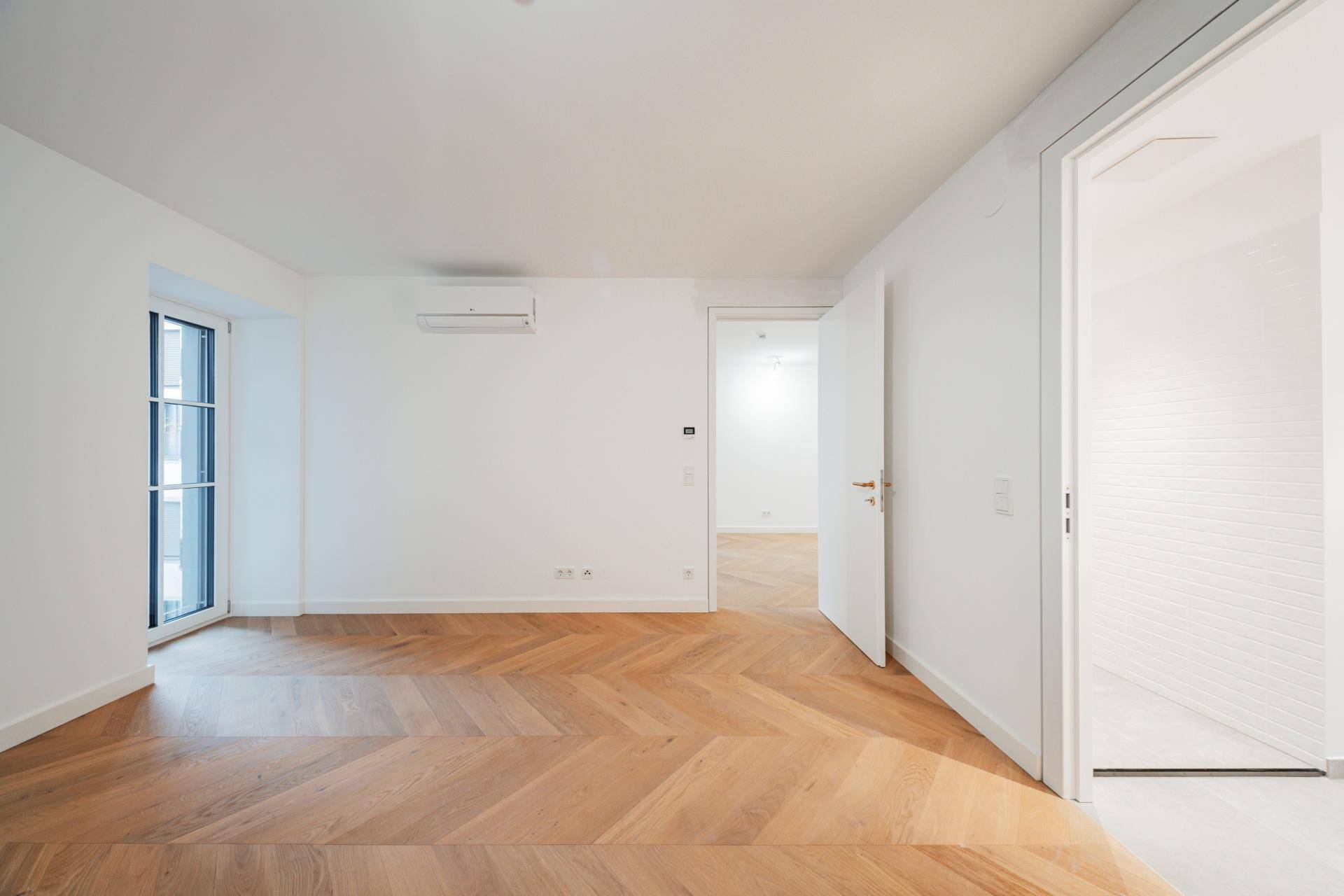 Heller Raum mit Fischgrätparkett, Klimaanlage und Zugang zu einem weiteren Zimmer.