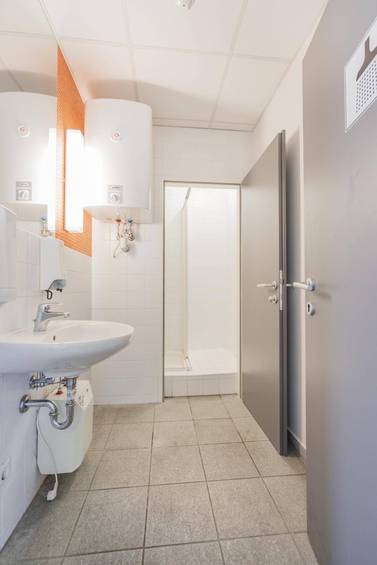 Öffentliche Toilette mit Waschbecken, Duschkabine und Warmwasserbereitern, hell gefliest.