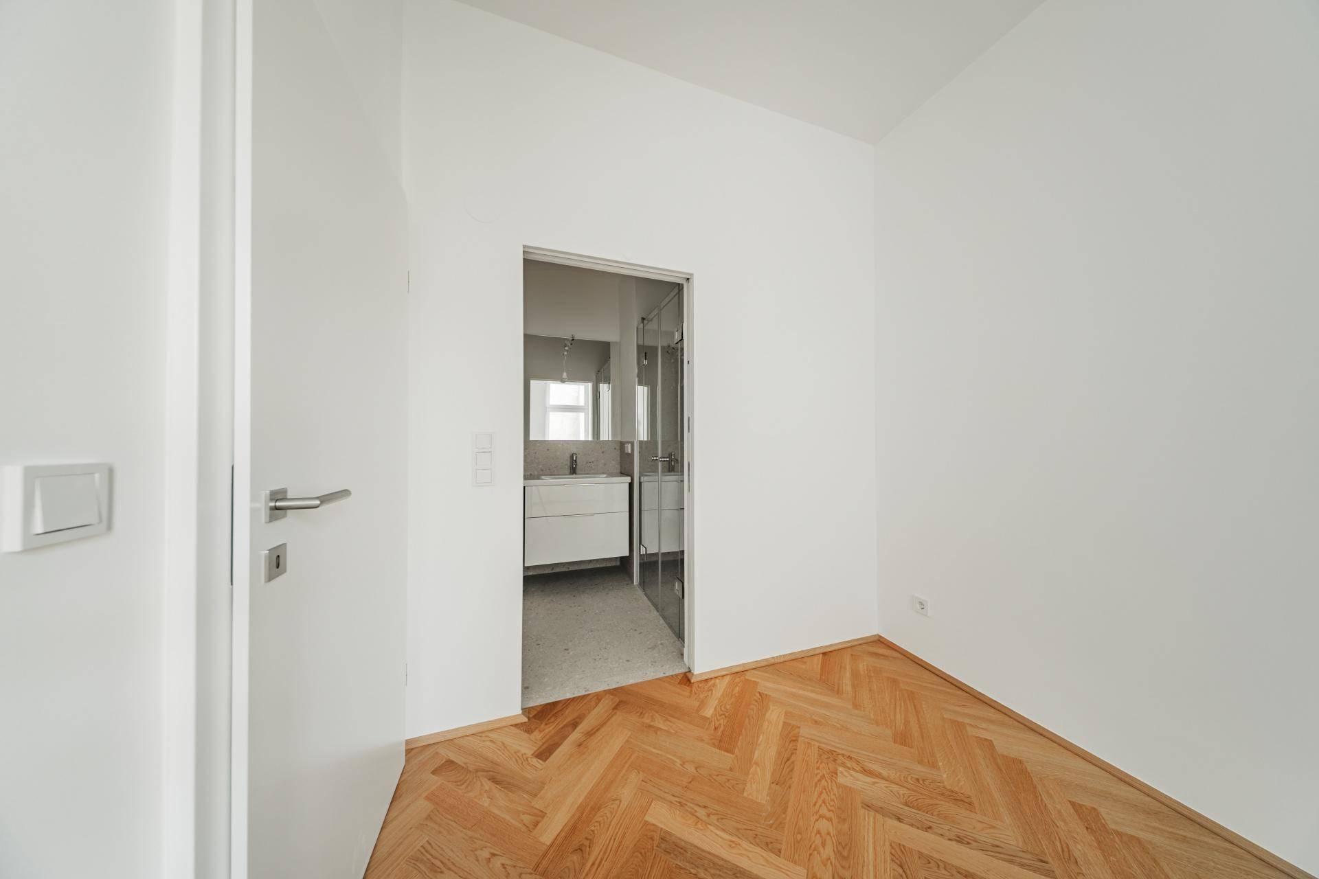 Heller Raum mit Fischgrätparkett und Zugang zu einem modernen Badezimmer mit Glasdusche.