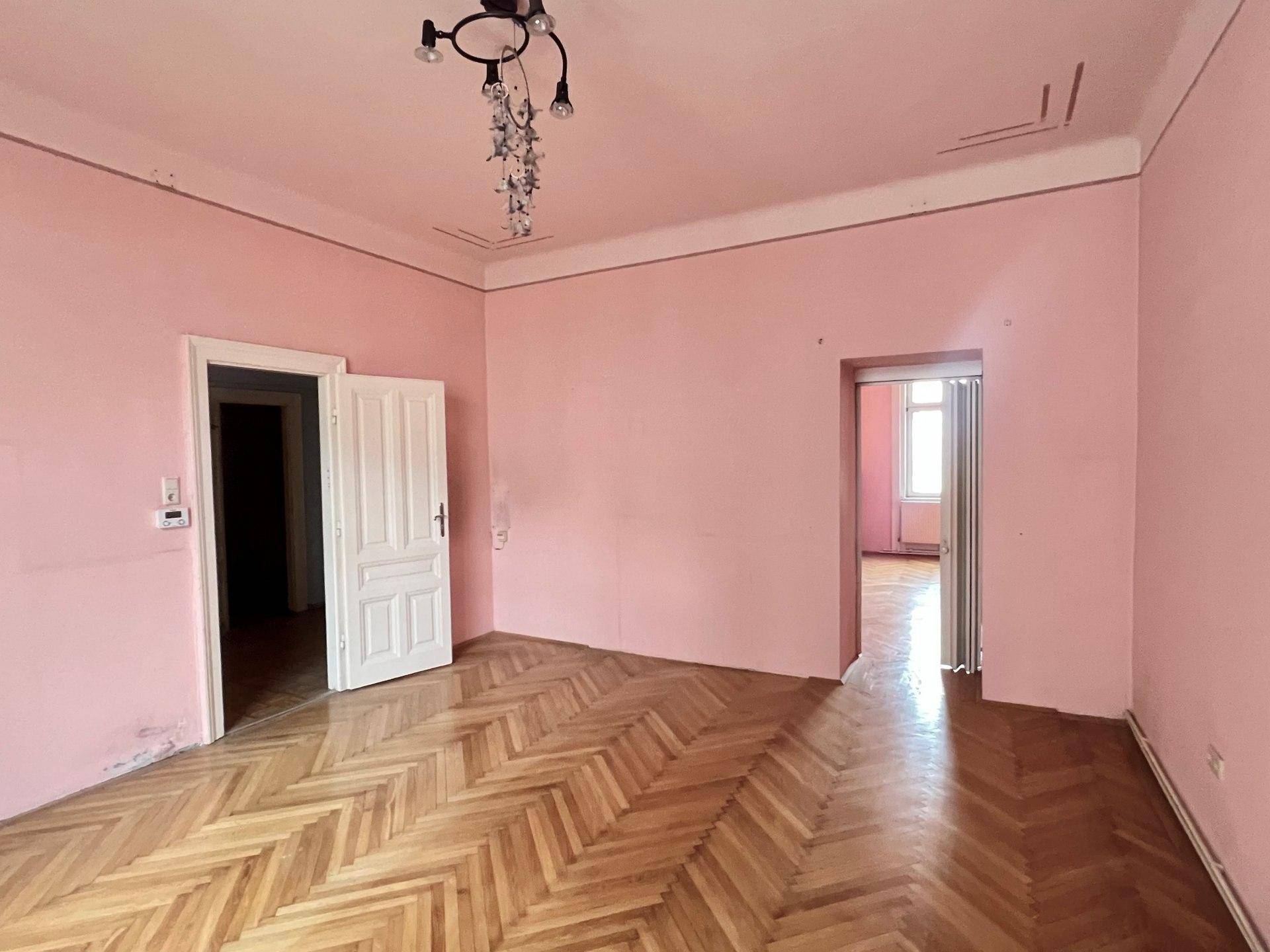 Großes Zimmer mit Fischgrätparkett, rosa Wänden und hohen Decken, mit Zugang zu weiteren Räumen.