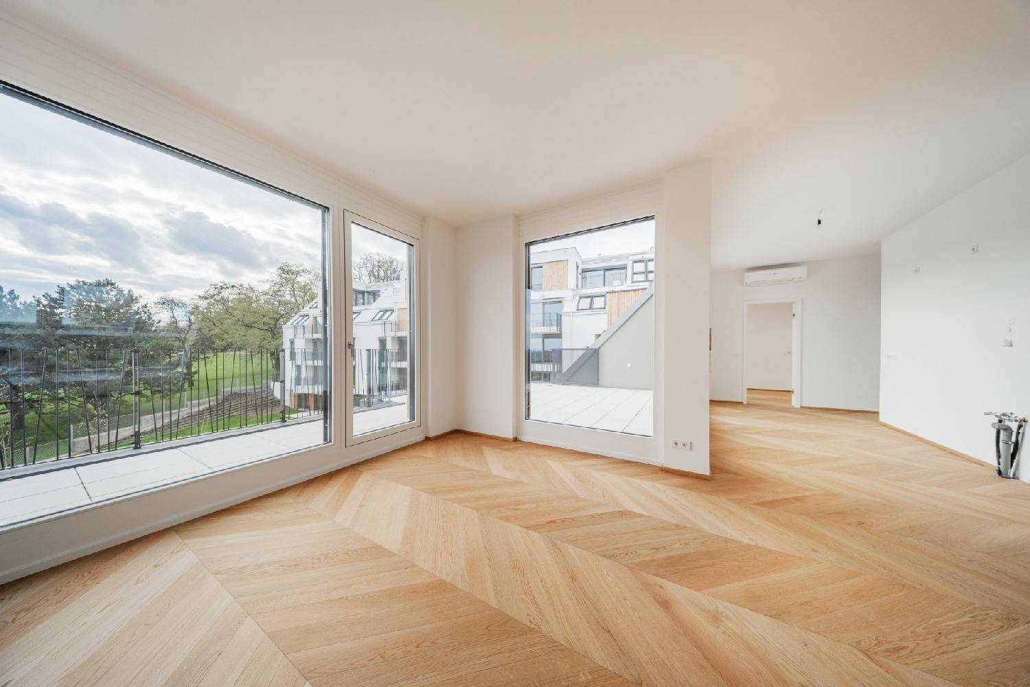 Helles, leeres Zimmer mit Fischgrätparkett und großen Fenstern, die einen Blick ins Grüne bieten.