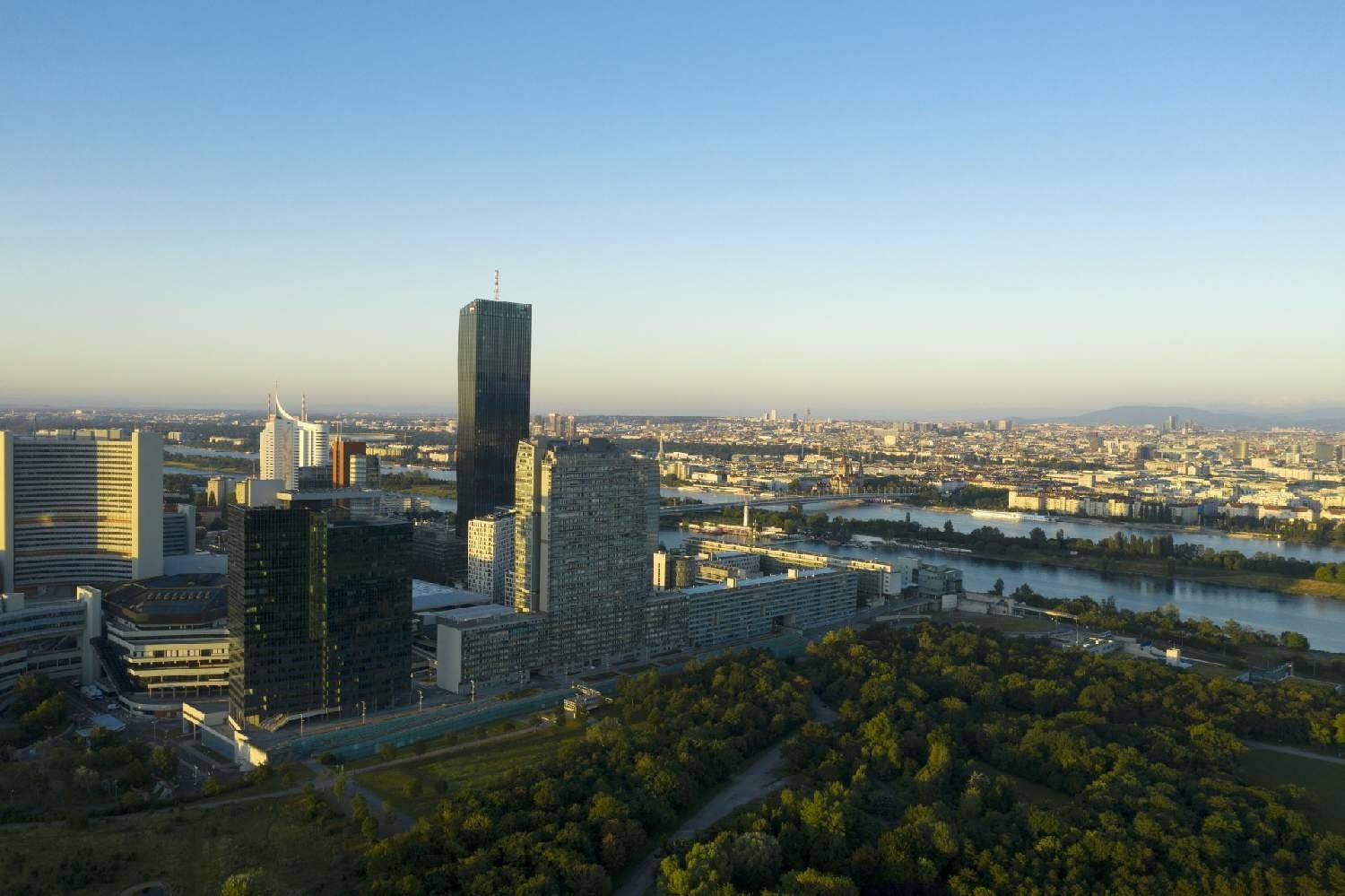 Panoramablick auf eine Stadt mit Fluss, grünen Flächen und markanten Hochhäusern bei Sonnenuntergang.