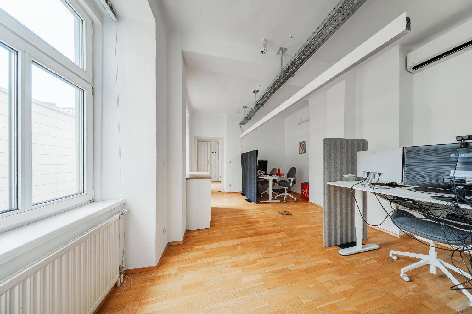 Langer Bürogang mit Holzboden, Fenstern und mehreren Arbeitsplätzen, gut beleuchtet.